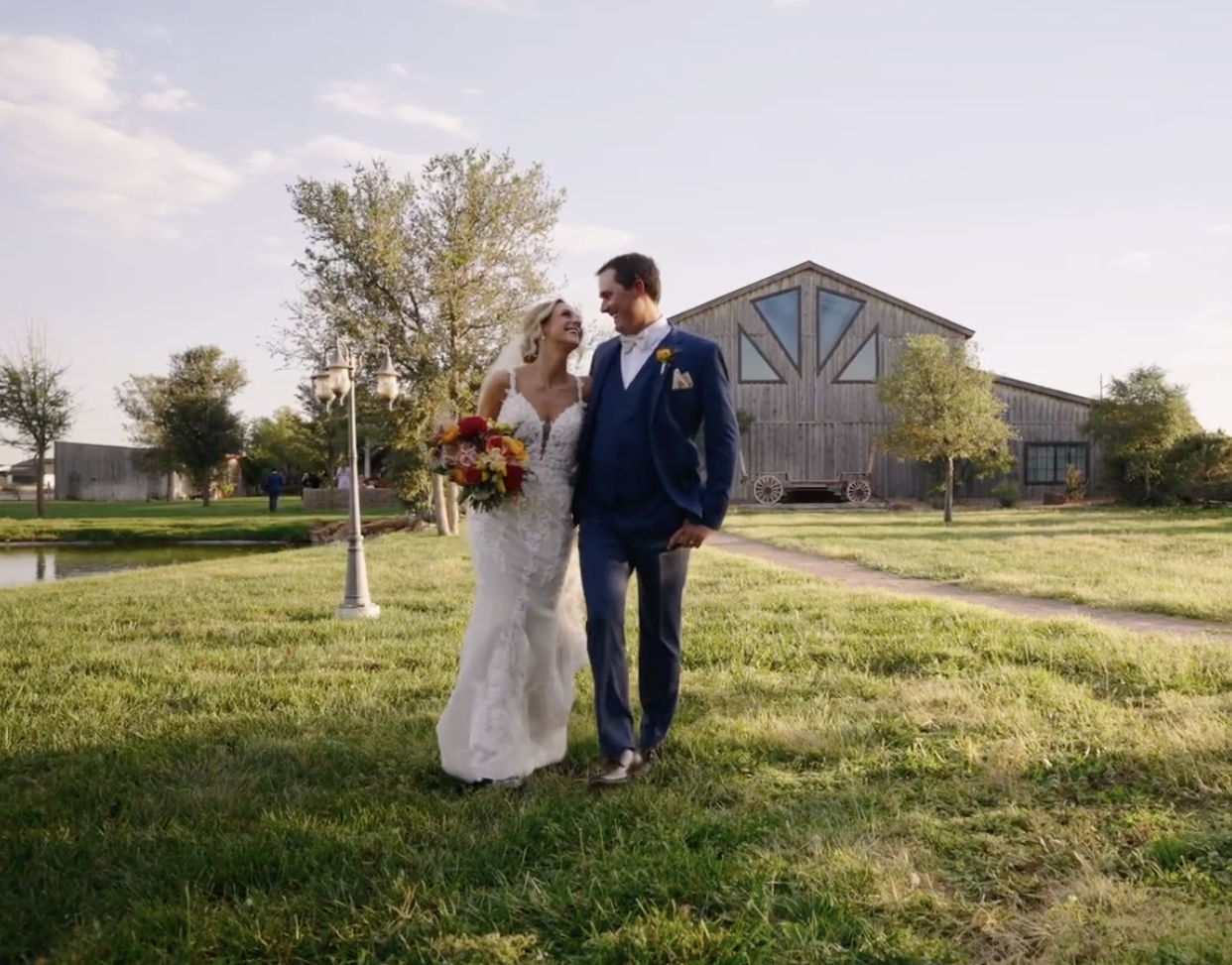 A newlywed couple walking outside on a sunny day, with a rustic barn in the background. The bride is holding a colorful bouquet and is smiling at the groom, who is dressed in a blue suit. They are on a grassy area with trees and pathway.