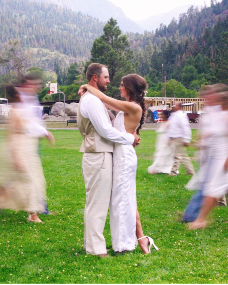 Couple dancing outdoors on green grass with blurred people dancing around them, mountains and trees in the background.