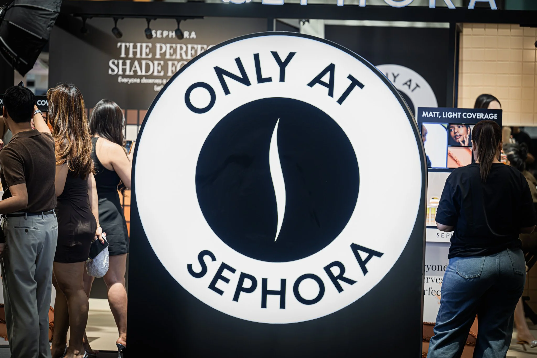 People standing near a large circular sign with the words 'ONLY AT Sephora' in black and white, in a busy retail environment.  Event photography.