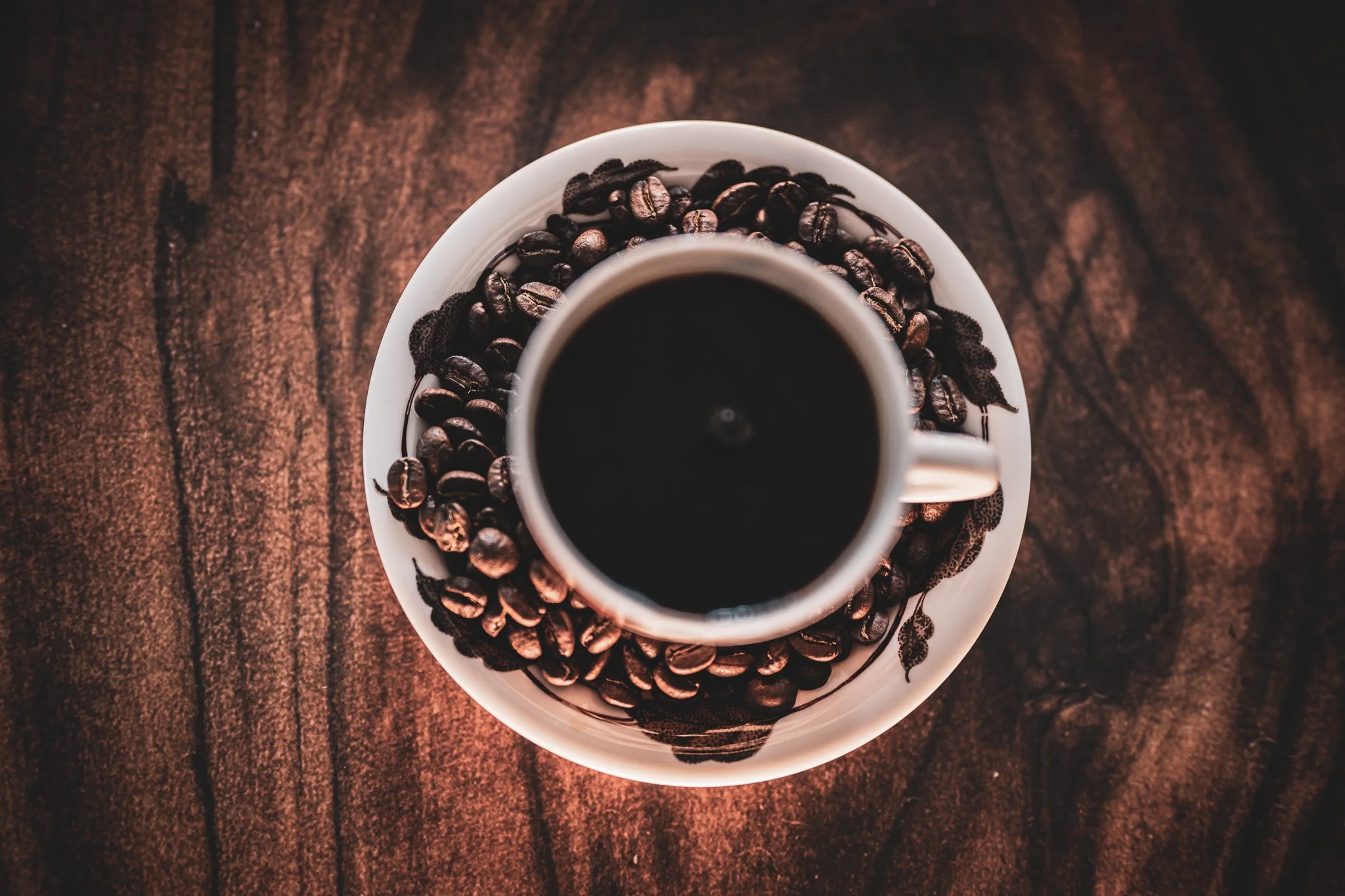 Top-down view of a white coffee cup filled with black coffee, placed on a matching saucer surrounded by roasted coffee beans on a dark wooden surface.