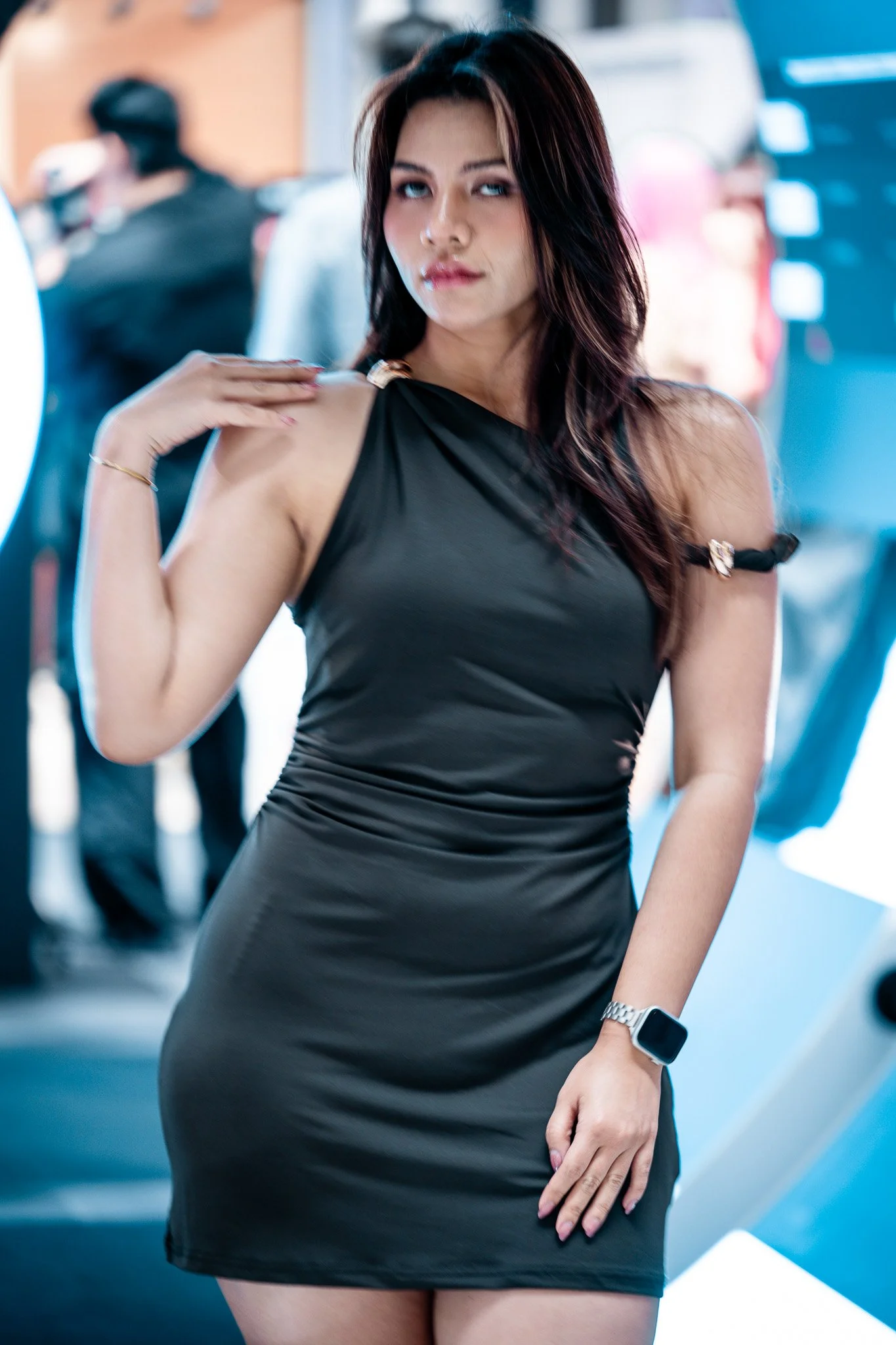 A woman with dark hair wearing a black dress with shoulder cutouts, posing in a modern, brightly lit indoor setting. She has a smartwatch on her wrist and a delicate bracelet, with a confident expression.  Event photography.