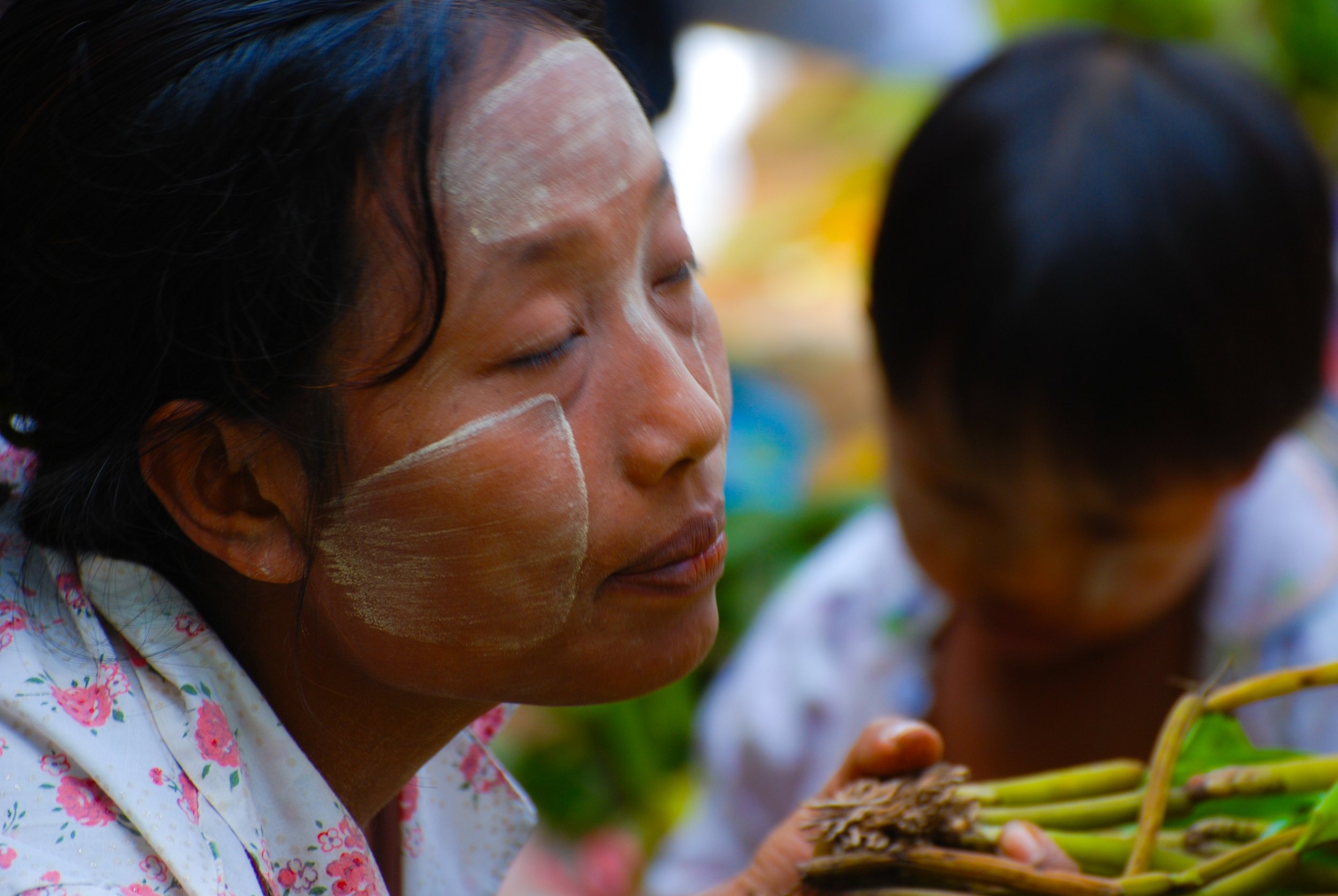 Close-up of a woman with traditional face paint, wearing a floral shirt, holding green plants with roots, outdoors.