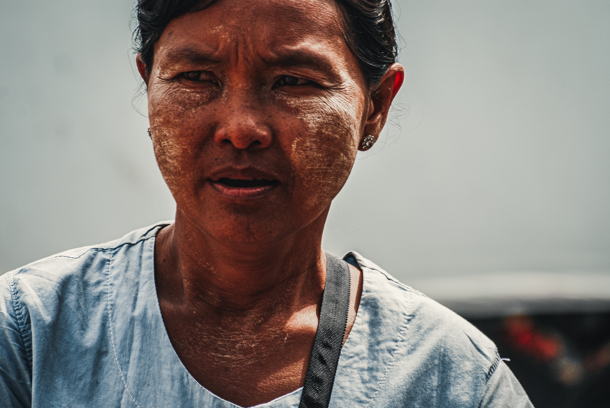 A close-up of a woman with short dark hair, earrings, and a distressed expression, wearing a light-colored shirt with a strap, against a plain background. Portrait photo