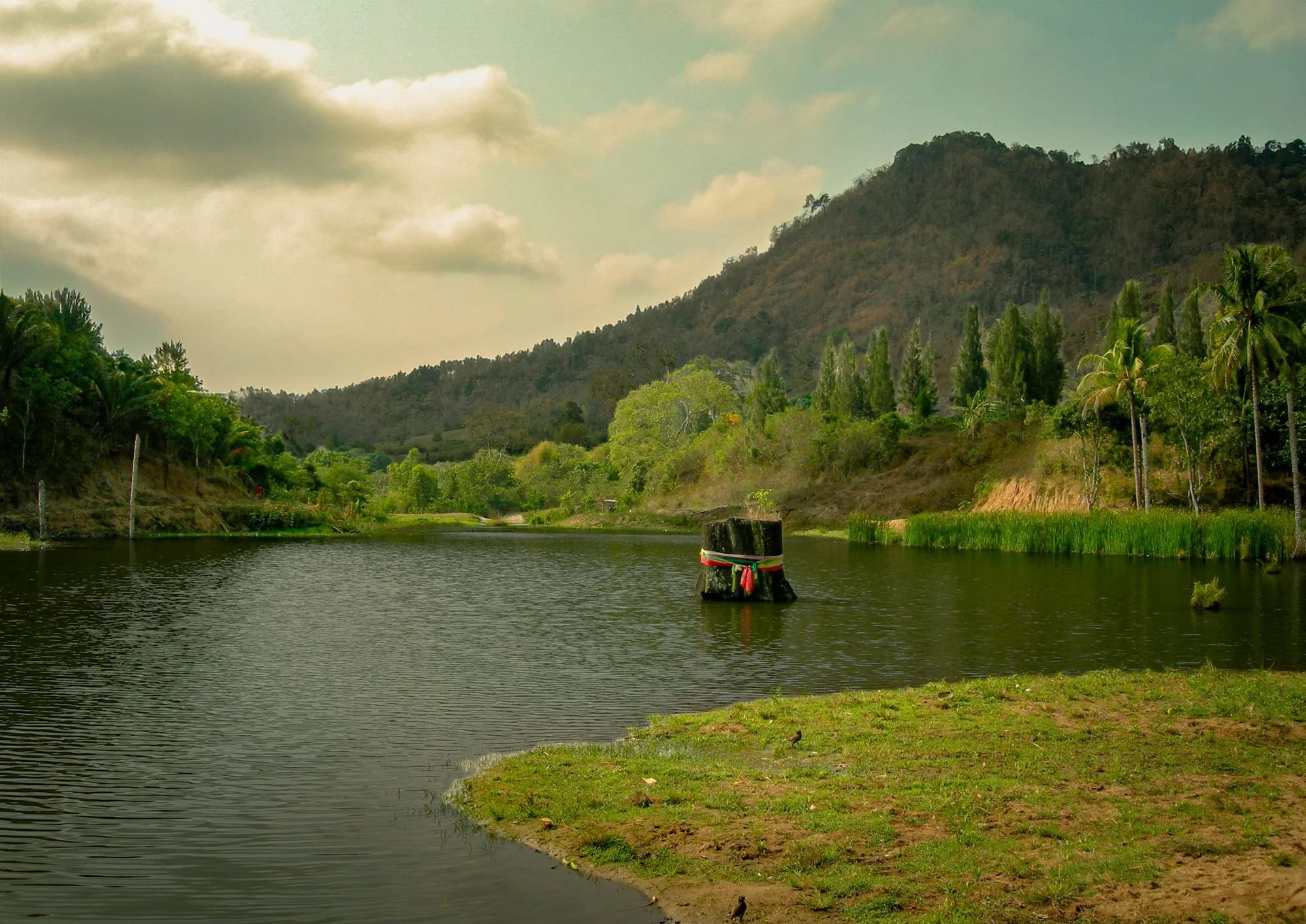 A river with green banks, trees, and a monument in the water, with a large hill in the background under cloudy skies.