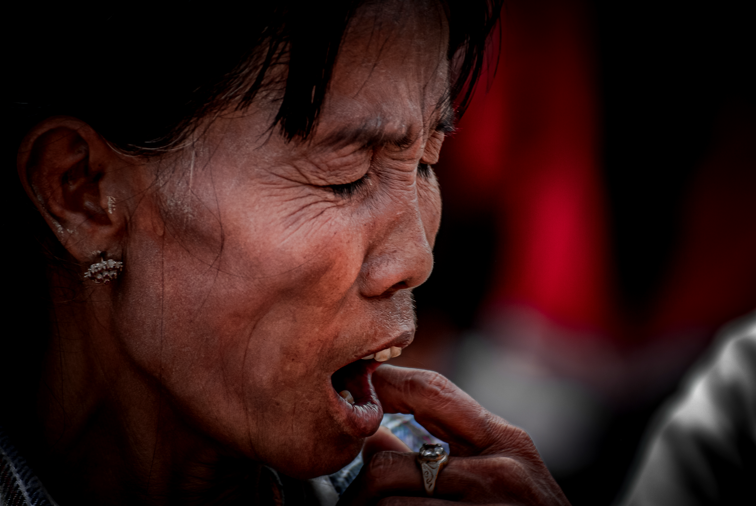 A close-up of a woman with dark hair, earrings, and wrinkles, smiling and touching her lips with her finger. Portrait photo.