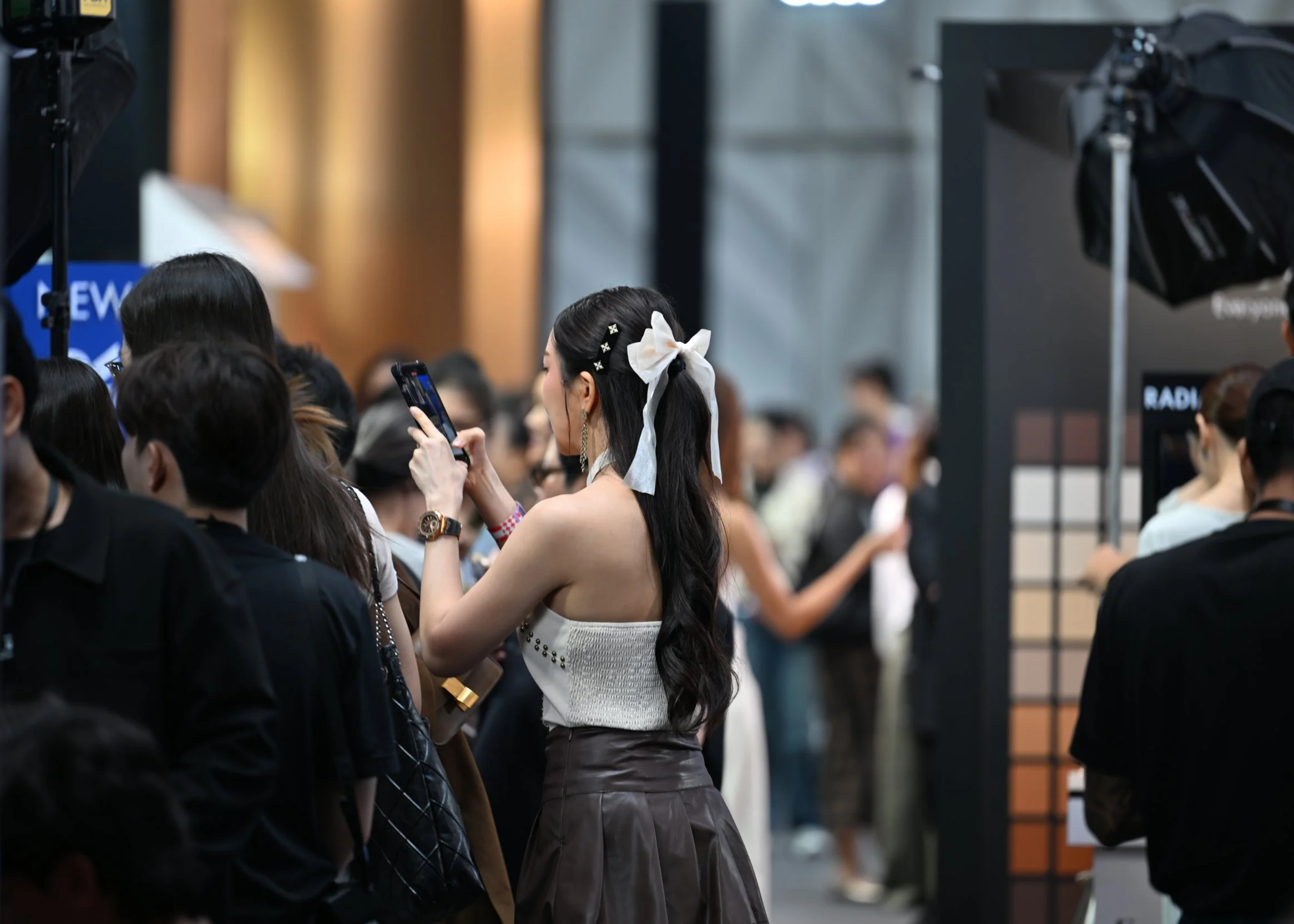 A woman with long dark hair, a white bow in her hair, and earrings, taking a photo with her smartphone at an indoor event or conference, surrounded by other attendees. Event photography.