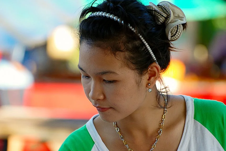 A woman with dark hair tied up, wearing a headband and earrings, looking down, with a blurred colorful outdoor background. Portrait photo