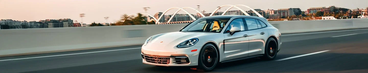 A silver luxury sports car drives on a highway with city buildings and a bridge in the background.