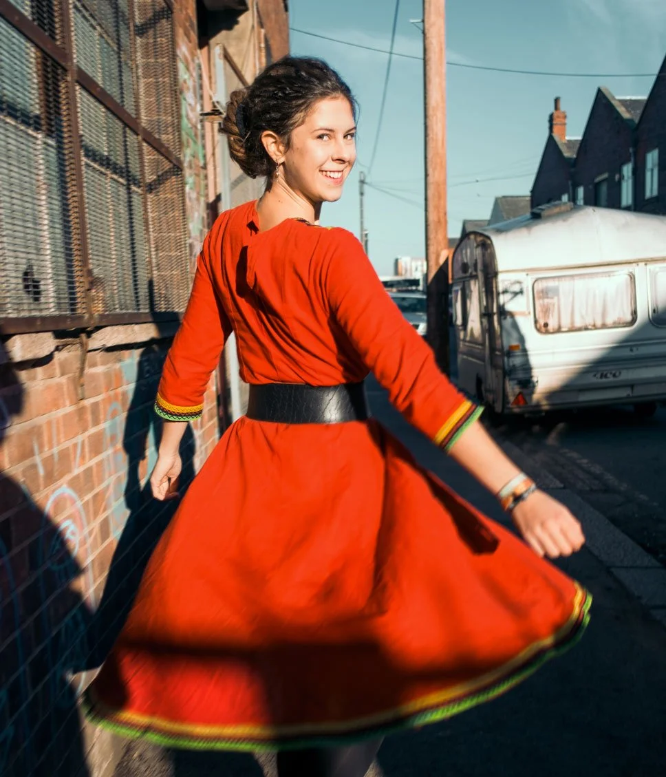 Eine lachende Frau in einem roten Kleid dreht sich und zeigt den Rücken, während sie draußen an einer Mauer mit einem Graffiti steht, mit Wohnhäusern und einem Wohnwagen im Hintergrund. Es ist sonnig und es herrscht eine angenehme, lebendige Atmosphä