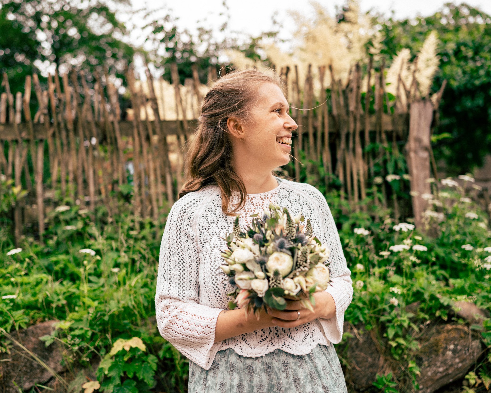 Frau im weißen Spitzenoberteil hält einen Blumenstrauß und lächelt im Garten bei Sonnenuntergang.