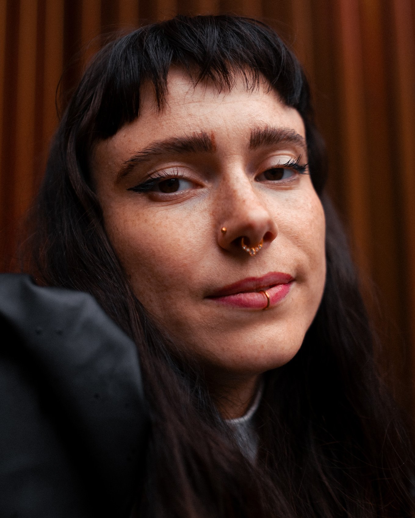 Nahaufnahme eines Menschen mit dunklen Haaren, mehreren Piercings und dezentem Make-up, vor einem Holz-Hintergrund.