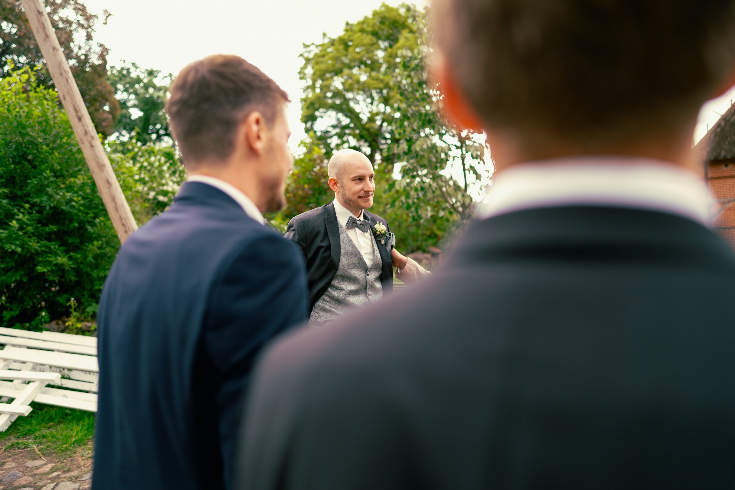 Ein Mann in formeller Kleidung, vermutlich bei einer Hochzeit, spricht zu zwei Männern, die ebenfalls formell gekleidet sind. Im Hintergrund sind Bäume und ein Holzmast zu sehen.