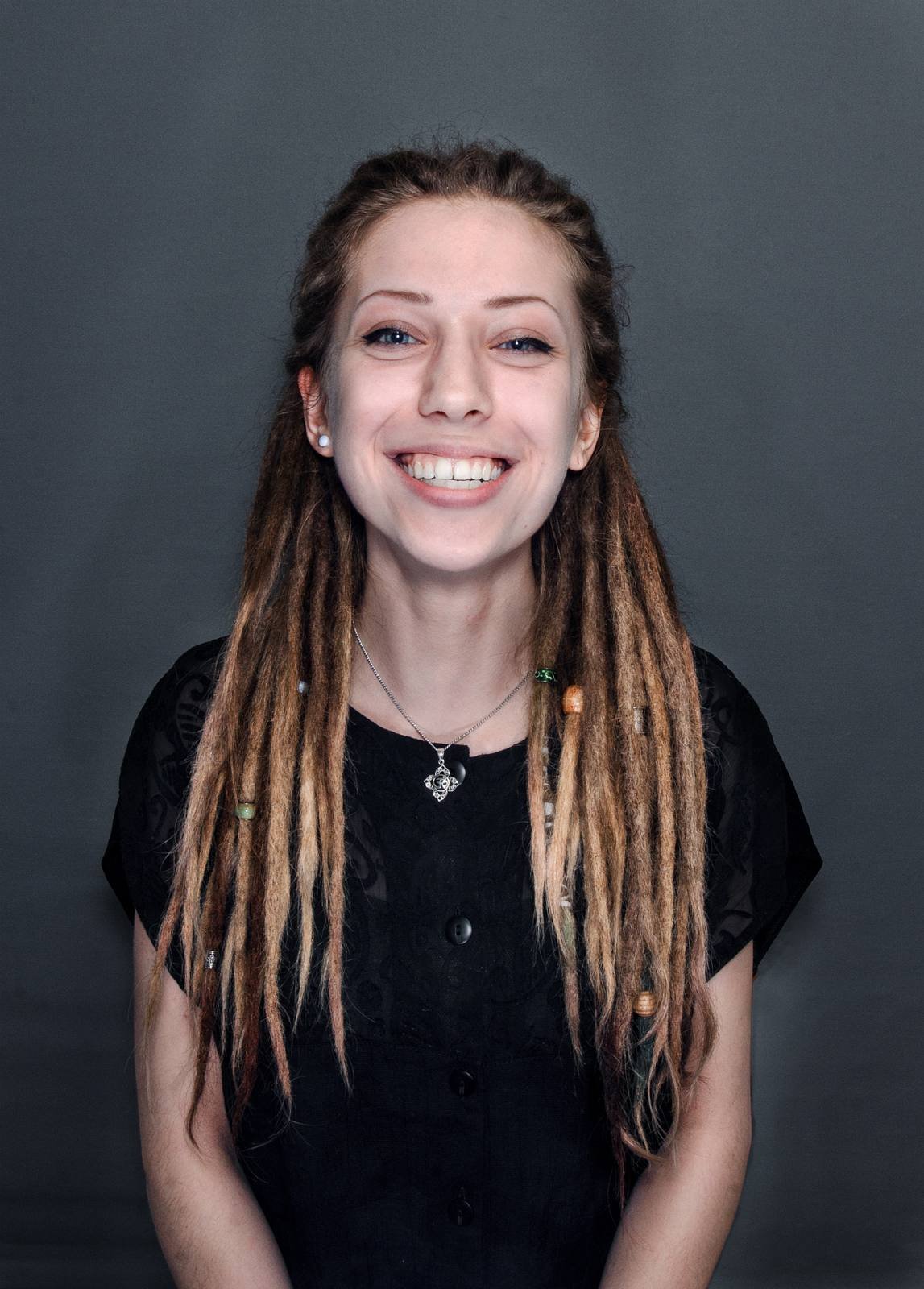 Lächelnde junge Frau mit Dreadlocks und schwarzen Kleidung vor grauem Hintergrund.