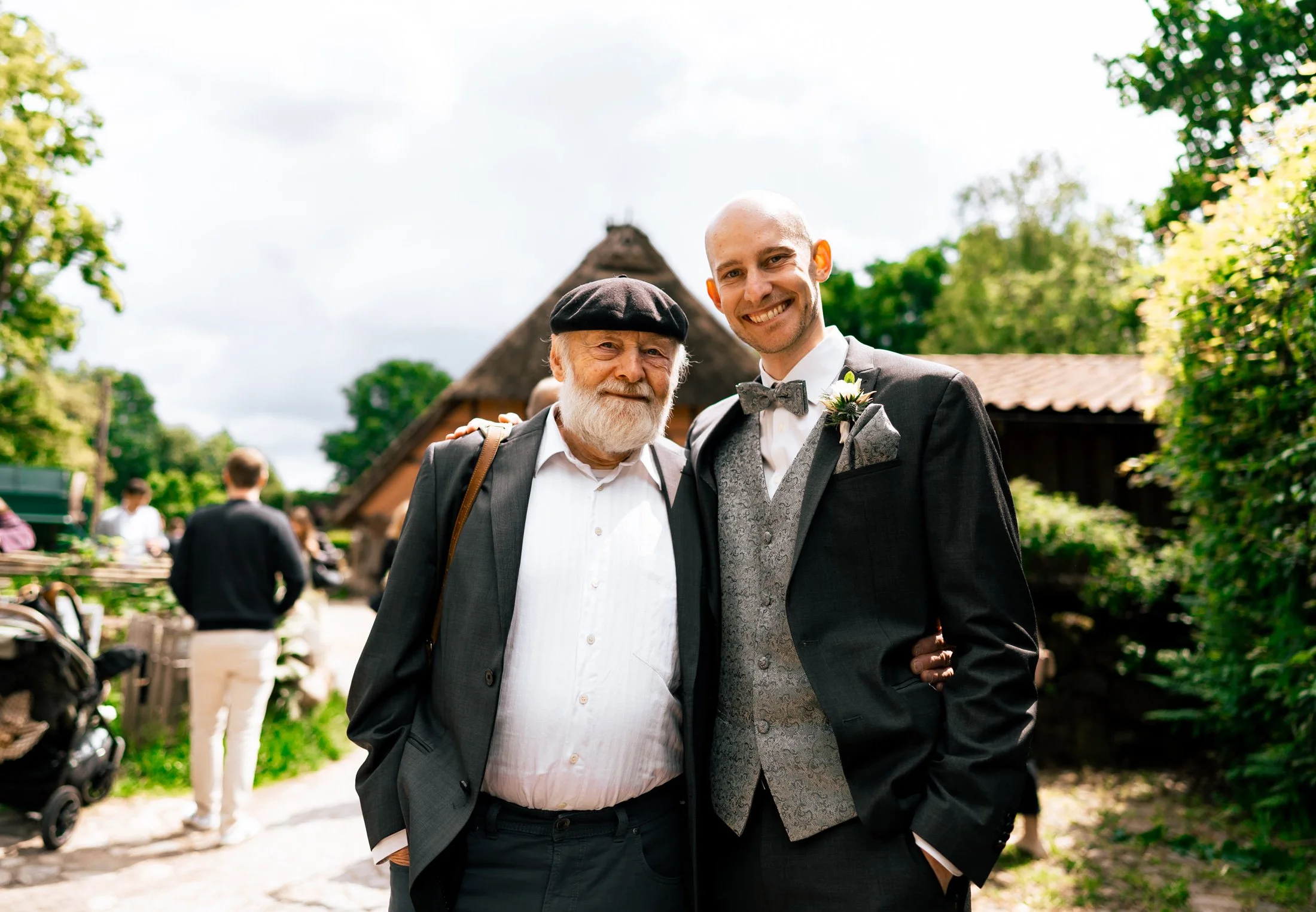 Zwei Männer, einer älter mit weißem Bart und einer jüngeren, beide in formeller Kleidung, stehen nebeneinander im Freien bei einer Hochzeit, umgeben von grünen Bäumen und traditionellen Gebäuden.