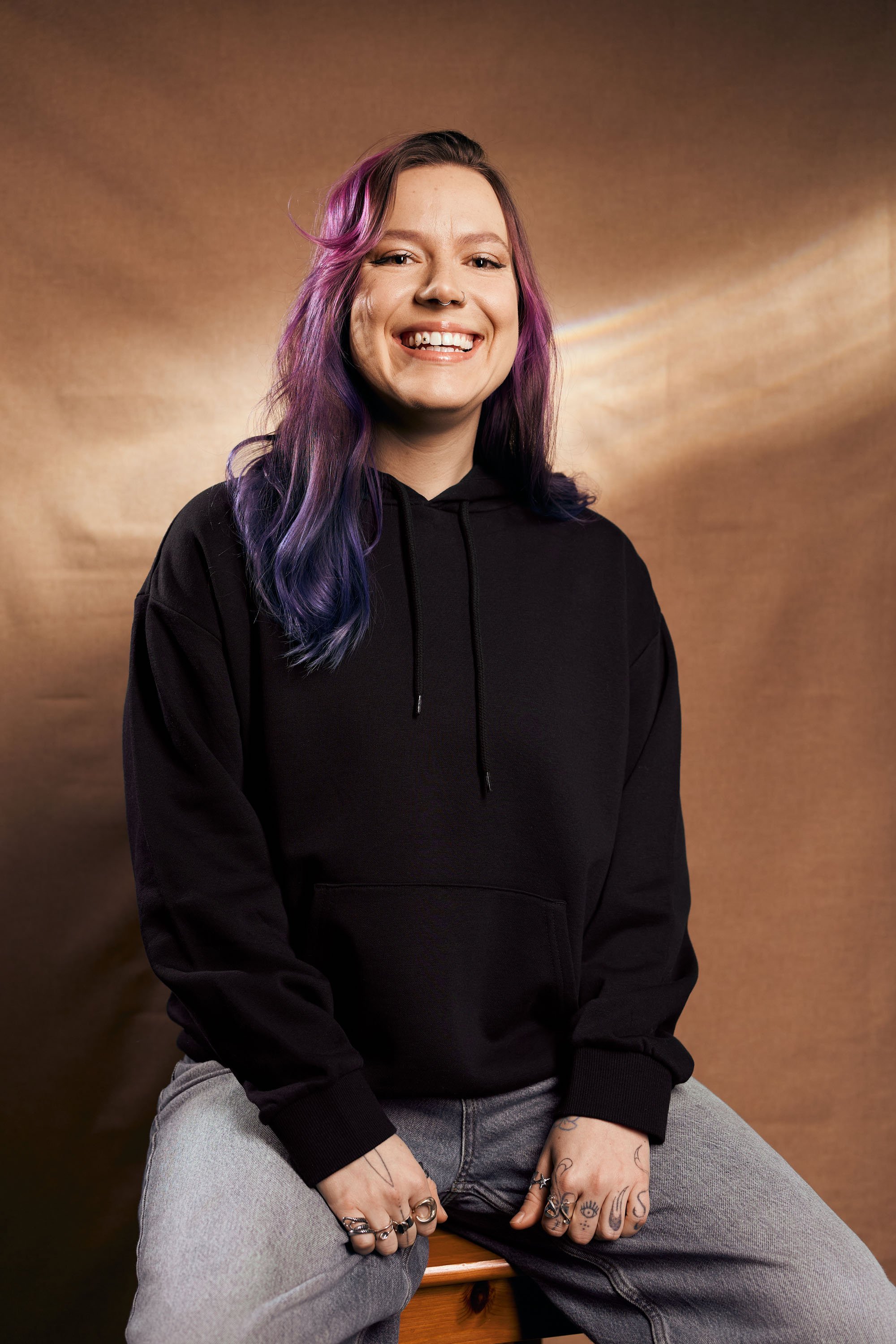 Junge Frau mit lila und blauer Haarfarbe, lächelnd, trägt schwarzen Hoodie und graue Jeans, sitzt auf einem Holzstuhl, Tattoos an den Händen, vor einem braunen Hintergund mit Lichtreflexen.