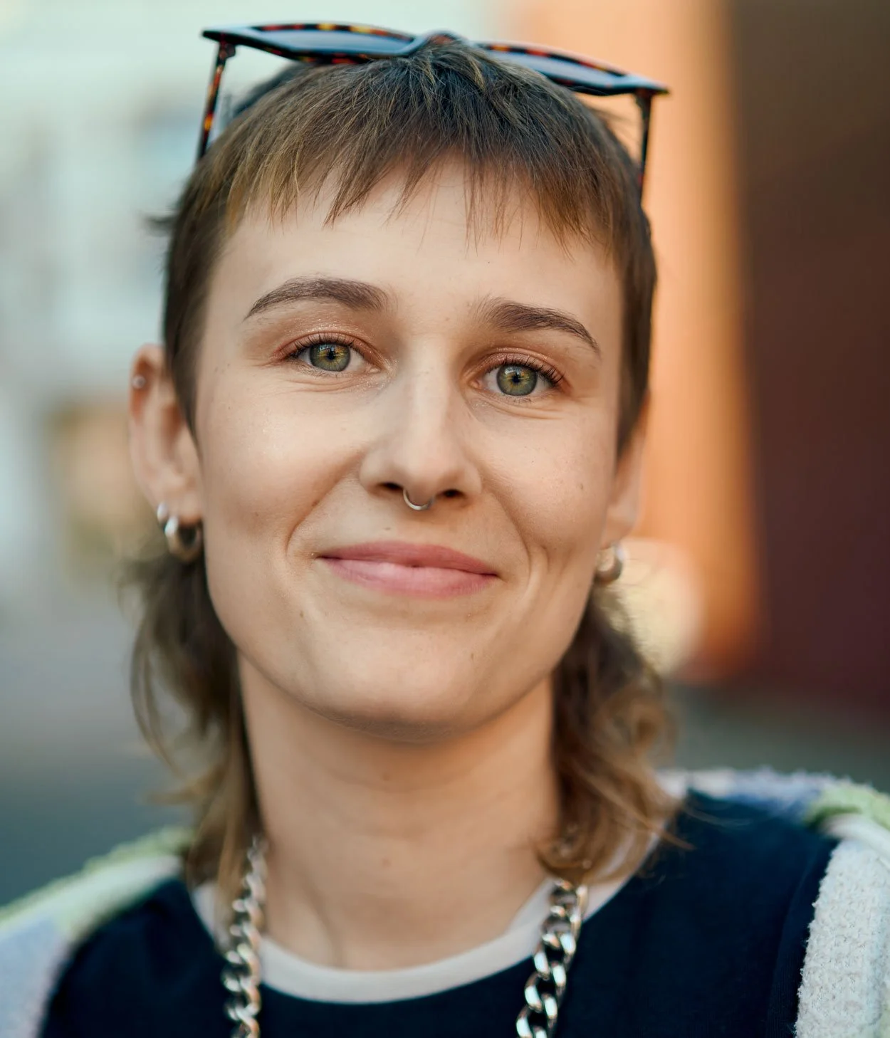 Nahaufnahme einer jungen Frau mit kurzen braunen Haaren, grünen Augen, dezentem Make-up, einem Nietenpiercing an der Nase, mehreren Ohrringen, einer Halskette und Sonnenbrille auf dem Kopf, im Freien bei Tageslicht.