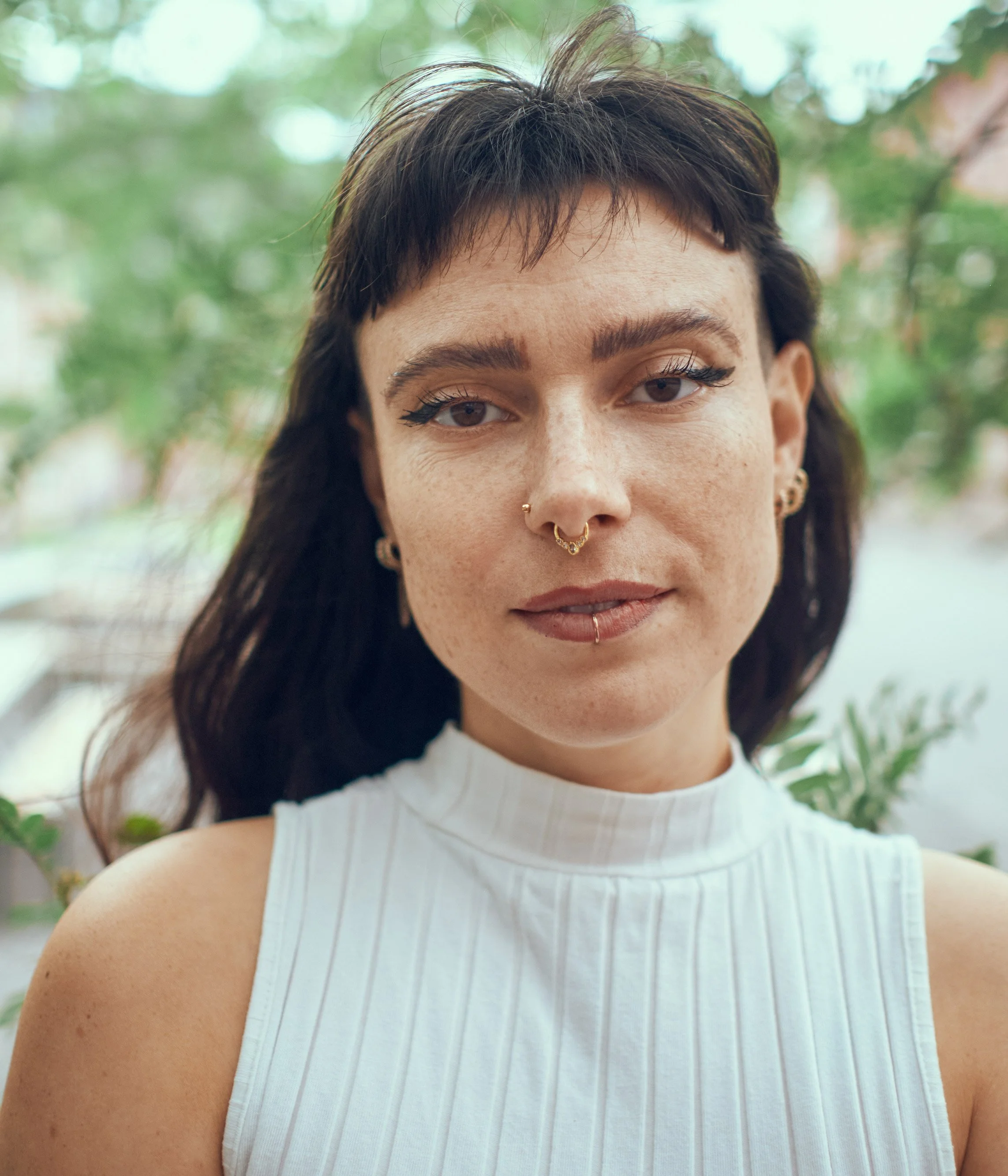 Nahaufnahme einer jungen Frau mit dunklen Haaren, mehreren Piercings im Gesicht, hellbraunen Augen und einem weißen ärmellosen Oberteil vor einem grünen, unscharfen Hintergrund.