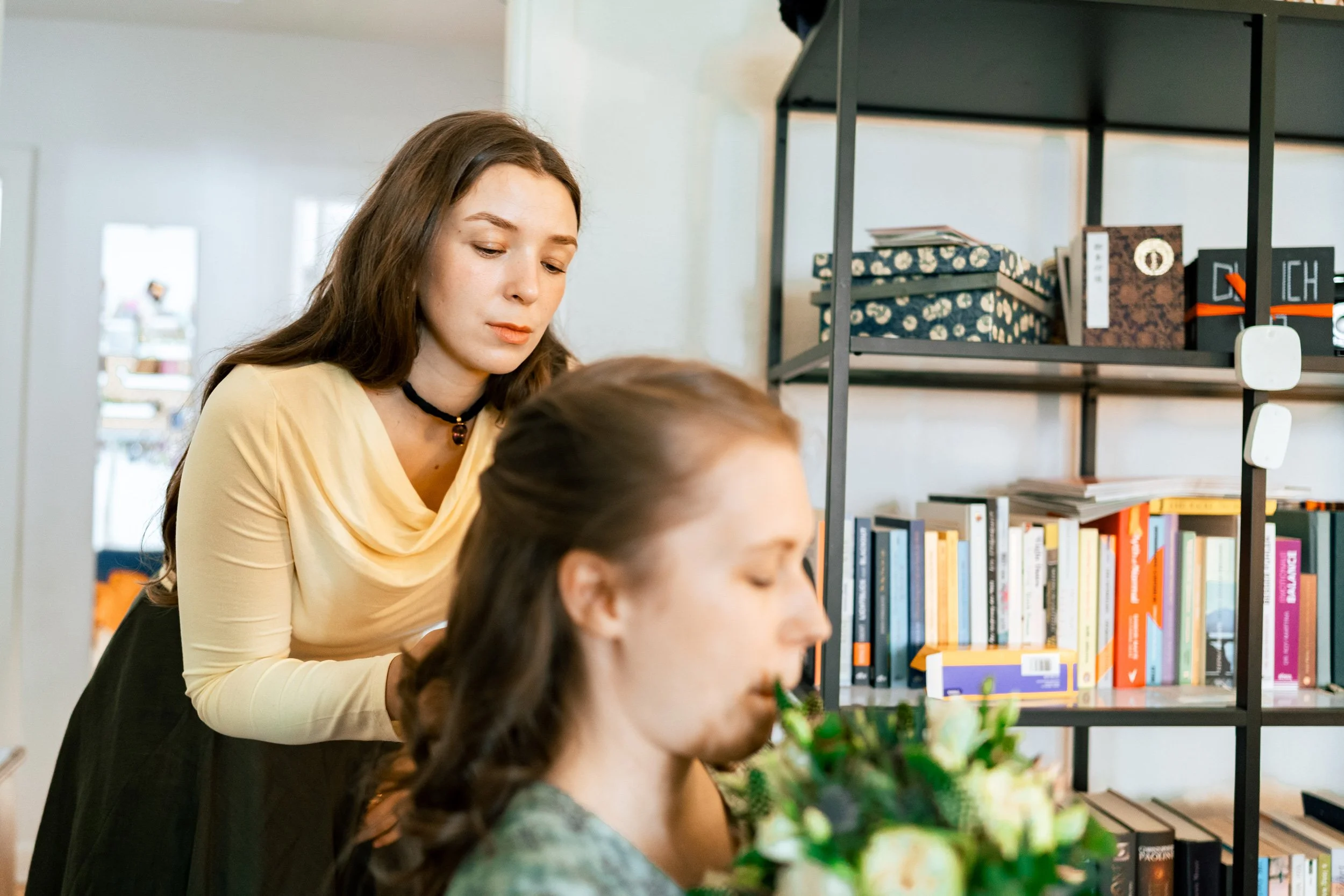 Zwei Frauen in einem Raum, eine sitzt mit geschlossenen Augen neben einem Blumenstrauß, die andere steht dahinter und schaut nach unten.