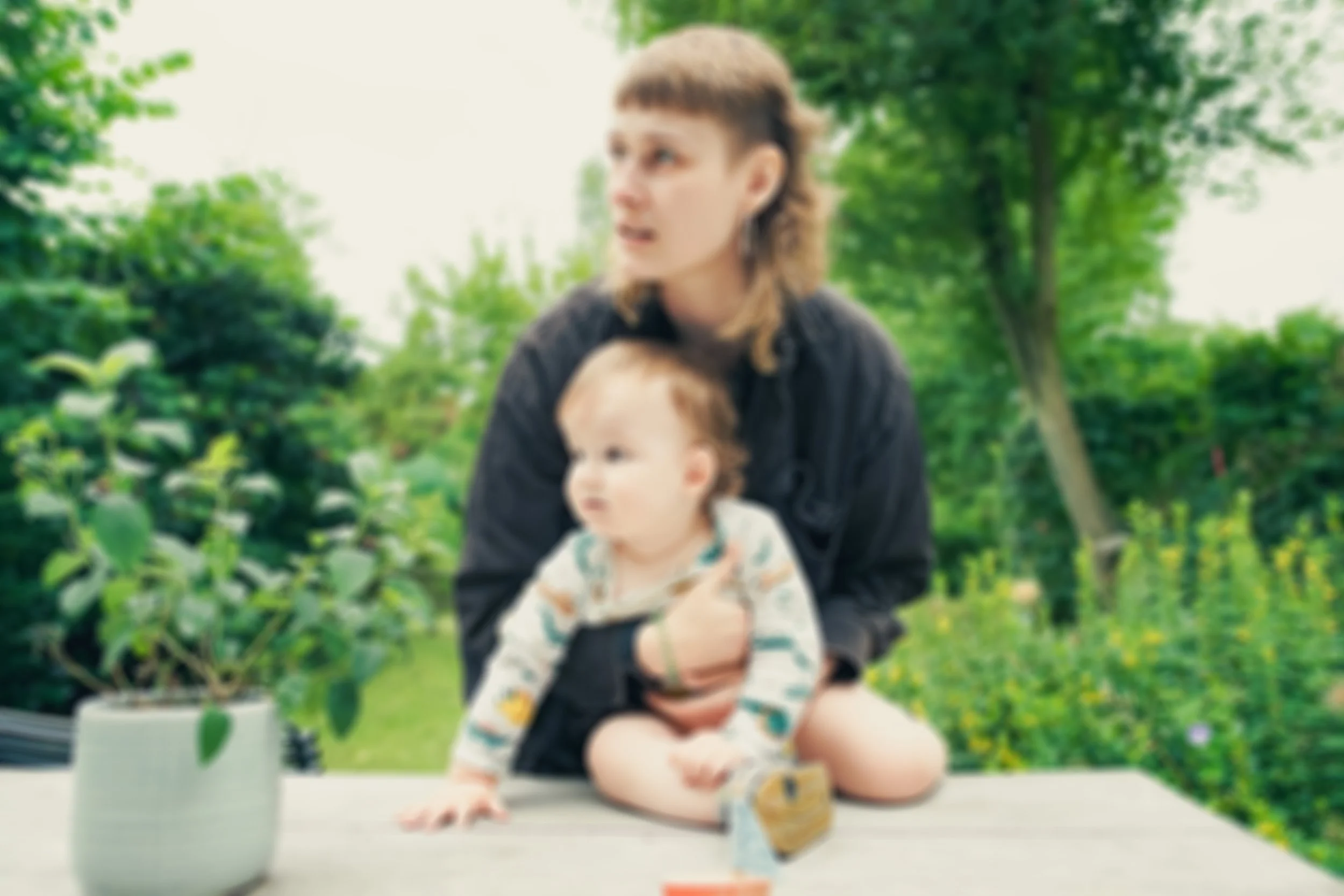 Eine Frau sitzt mit einem kleinen Kind im Garten, umgeben von grünen Bäumen und Pflanzen.