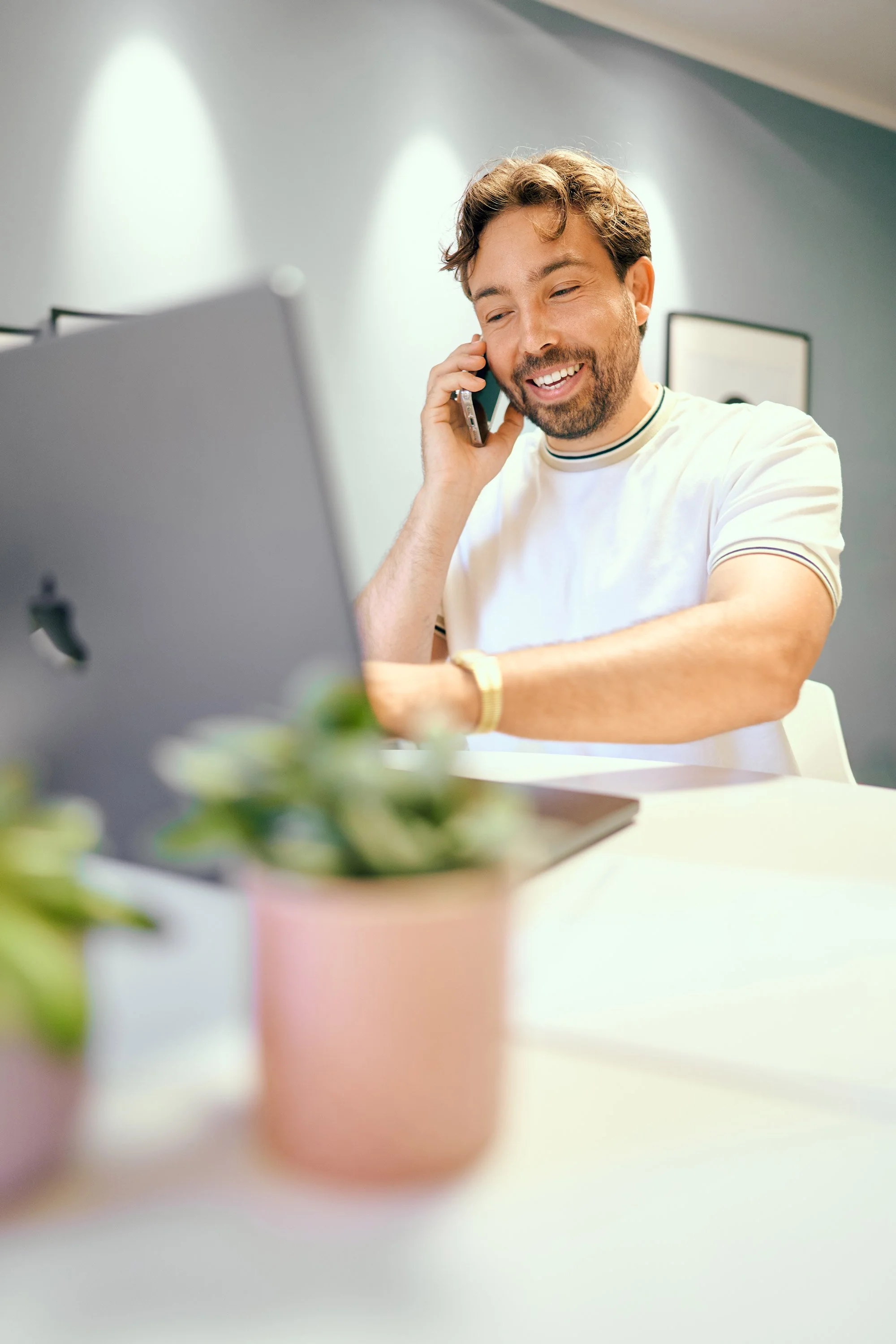 Mann sitzt an einem Tisch, spricht auf einem Handy, lächelt, neben einem Laptop, im Hintergrund eine Wand mit Bildern.