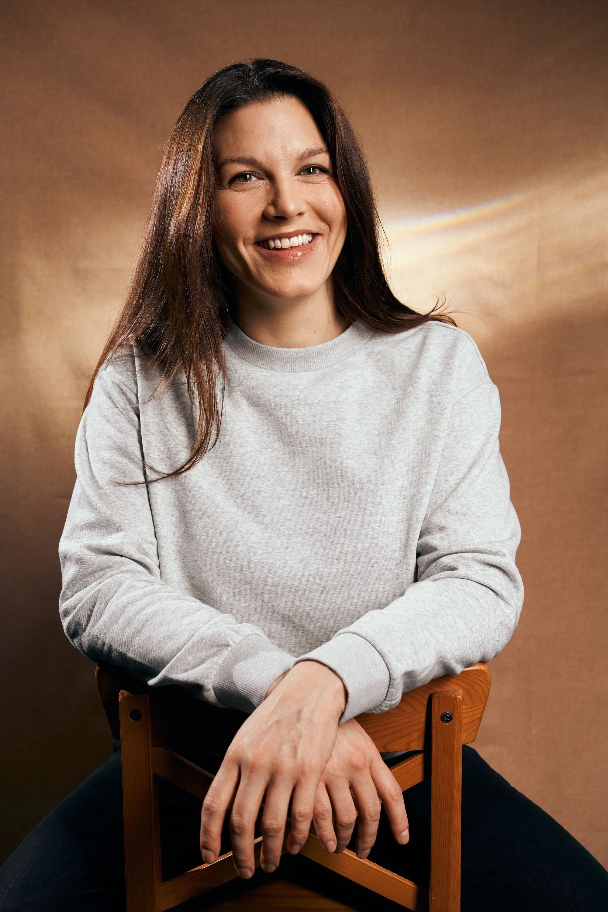 Junge Frau mit braunen Haaren, grauem Sweatshirt, lächelnd, sitzt auf einem Holzstuhl vor braunem Hintergrund.