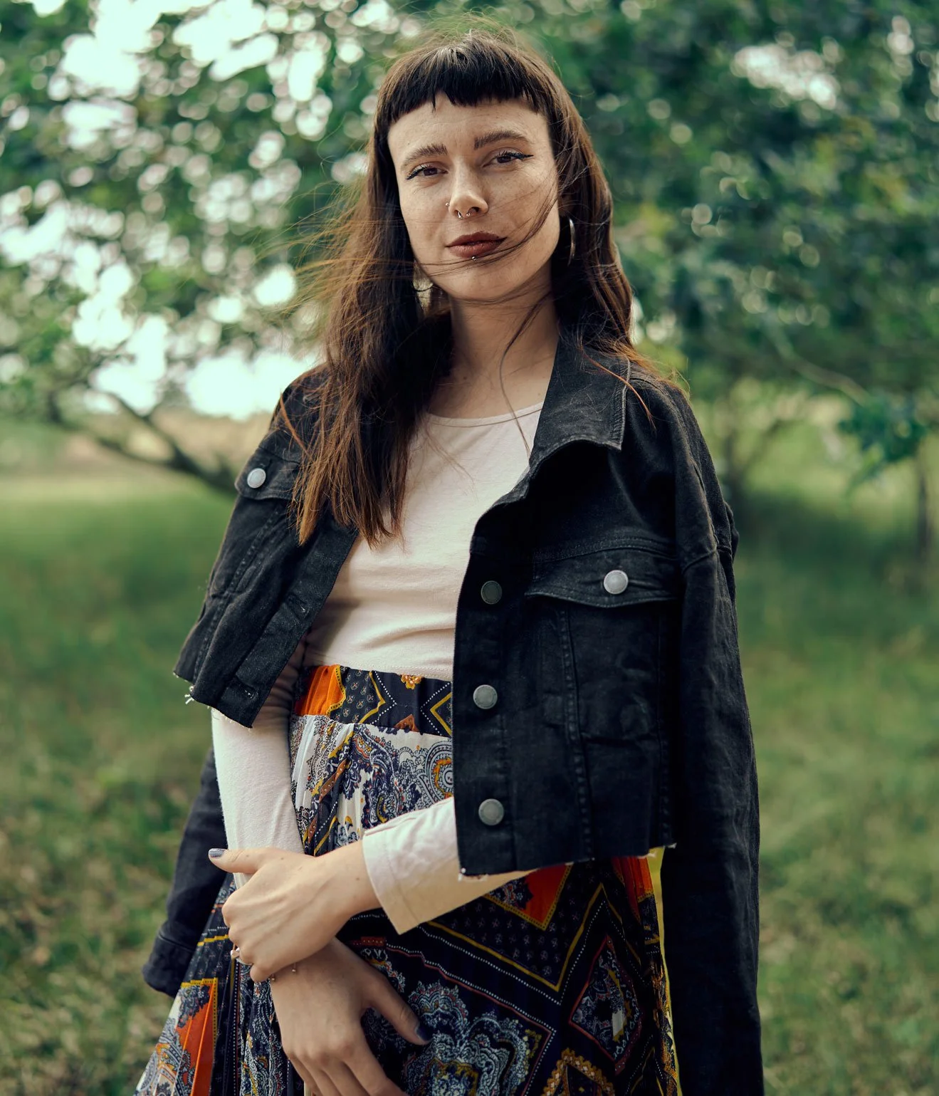 Eine junge Frau mit dunklem Haarschnitt und Piercings steht im Freien vor grünen Bäumen, trägt eine schwarze Jeansjacke, ein weißes Oberteil und einen bunten Rock.