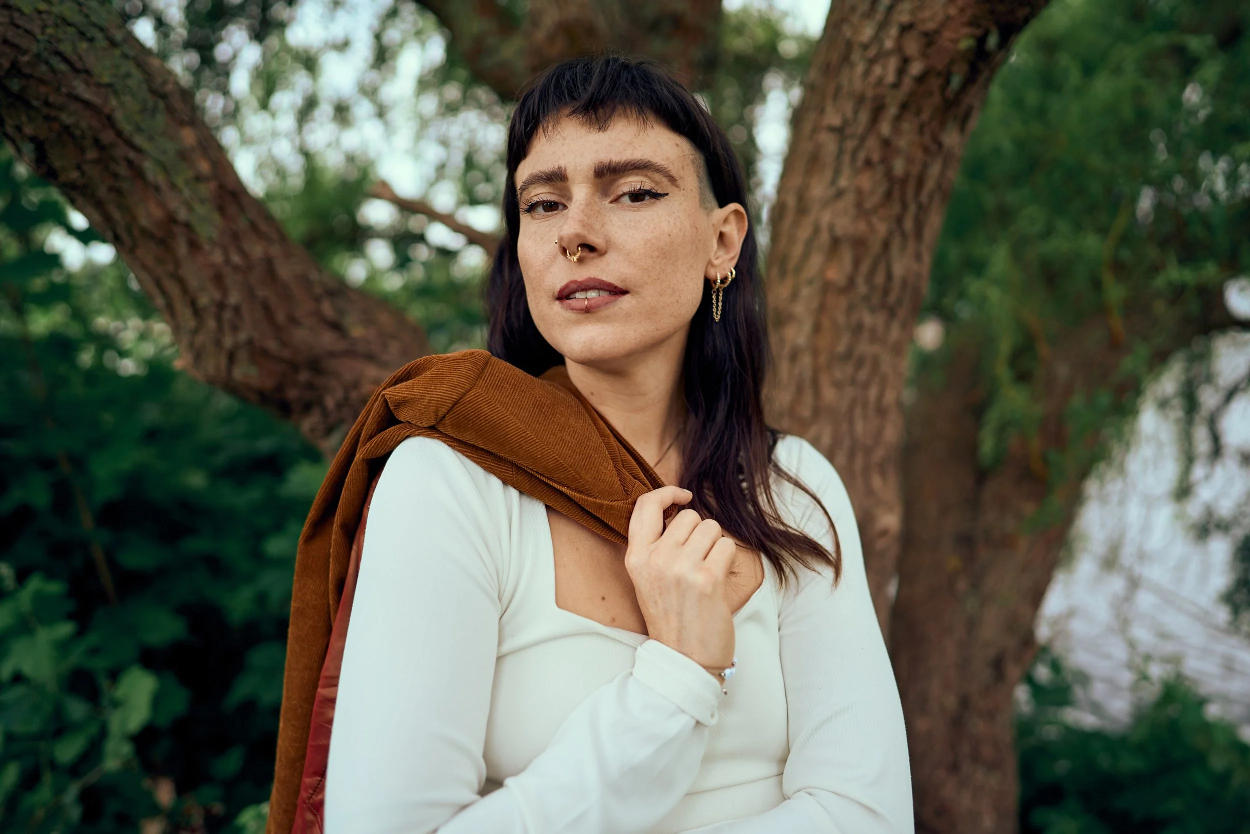 Eine Frau mit dunklem, kurzem Haar und Piercings, trägt einen weißen Pullover und einen braunen Rucksack, steht vor einem Baum im Grünen.