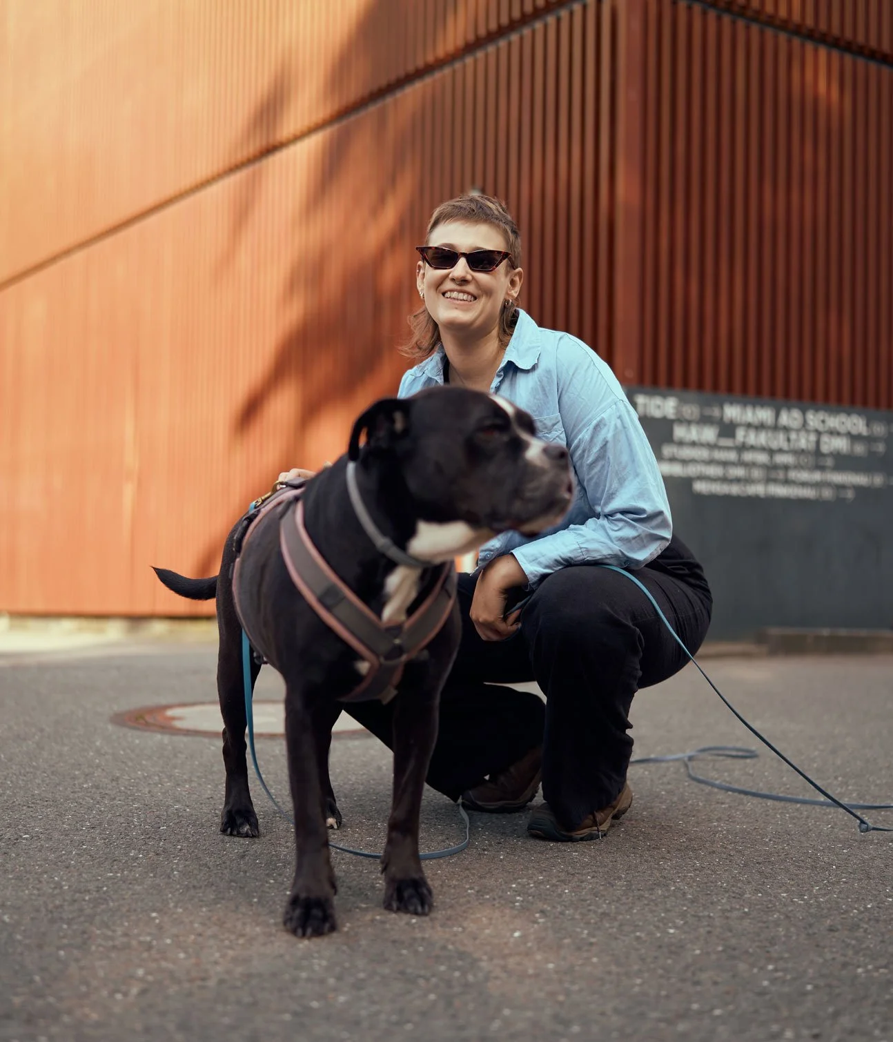 Eine lachende Frau mit Kurzhaarschnitt und Sonnenbrille ist draußen neben einem großen schwarzen Hund mit Knoten und Geschirr. Im Hintergrund ist ein modernes Gebäude mit einer Holzfassade und einem schwarzen Schild mit weißen Texten.