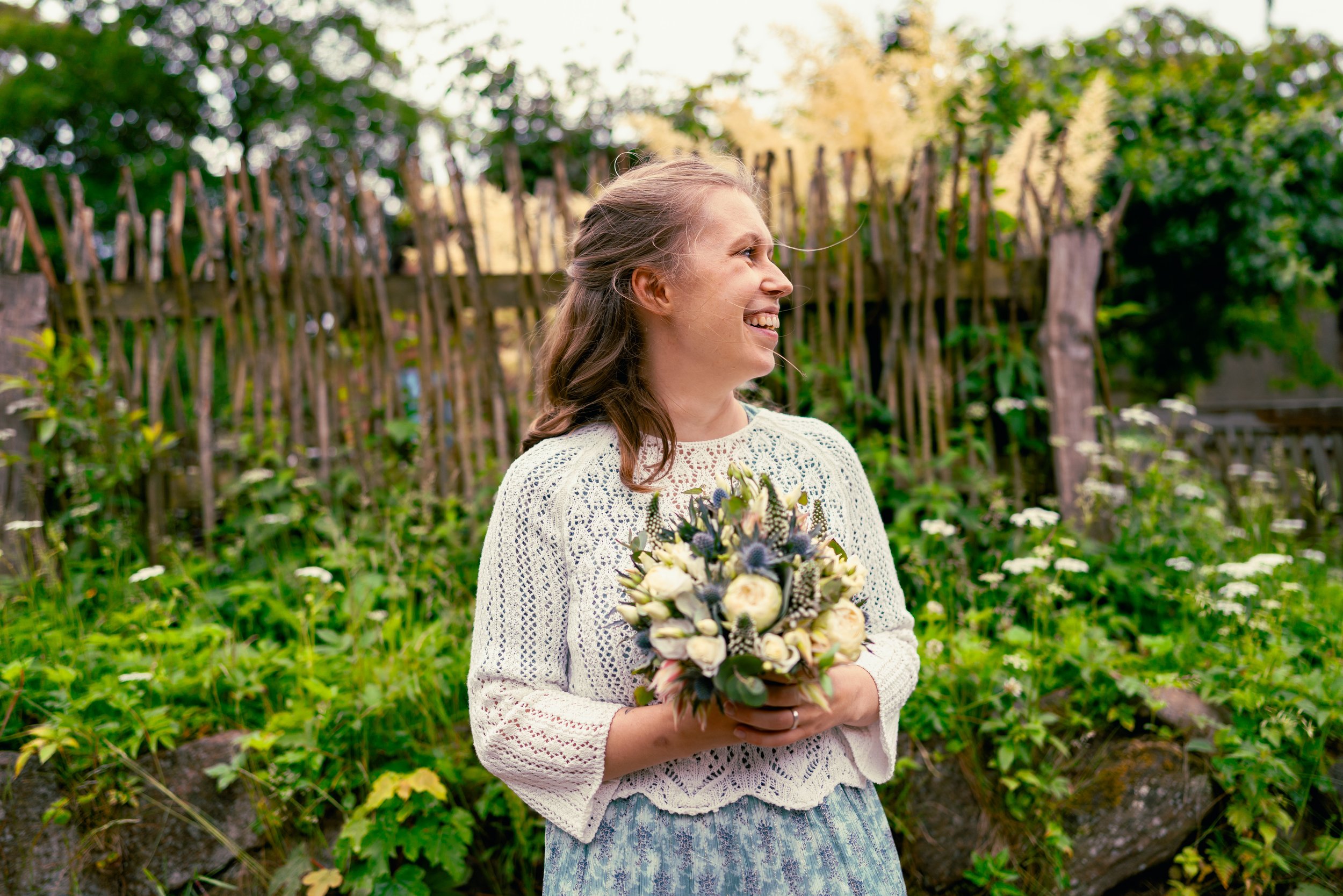 Junge Frau mit Blumenstrauß, lachend im Garten, umgeben von grünen Pflanzen und Holzhecke.