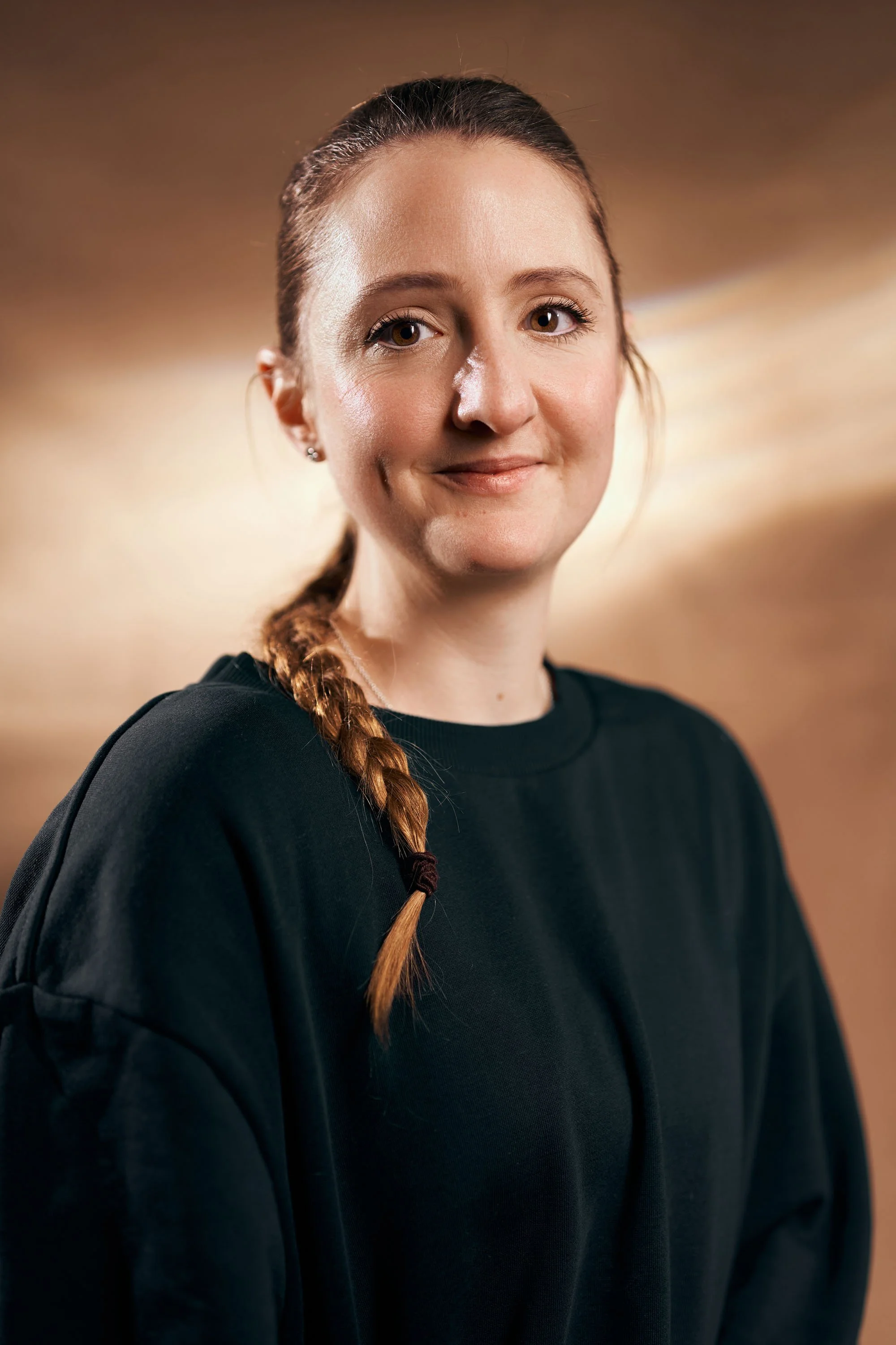 Junge Frau mit braunem Zopf in einem schwarzen Pullover, lächelnd, vor hellem, unscharfem Hintergrund.