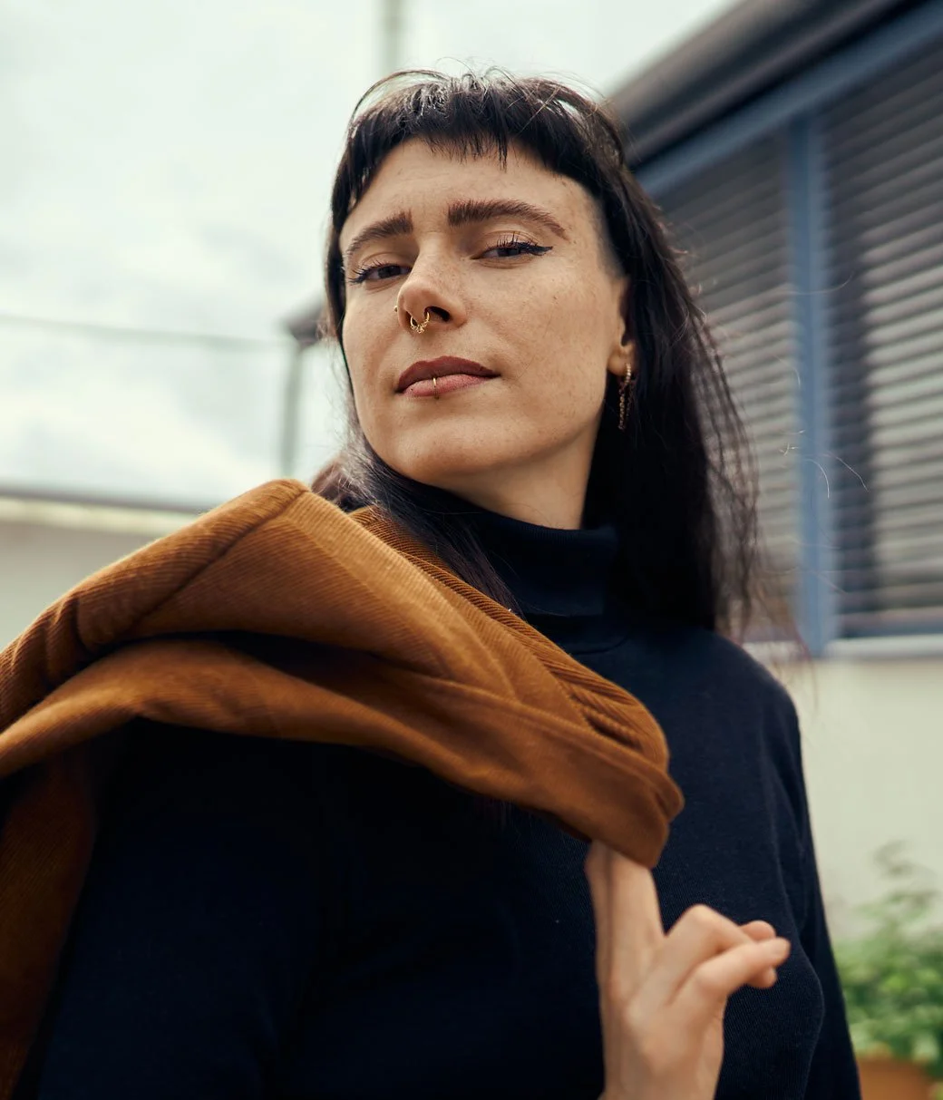 Eine junge Frau mit dunklen, kurzen Haaren trägt eine schwarze Kleidung und einen braunen Mantel über der Schulter, blickt selbstbewusst in die Kamera, mit einem Gebäude im Hintergrund.