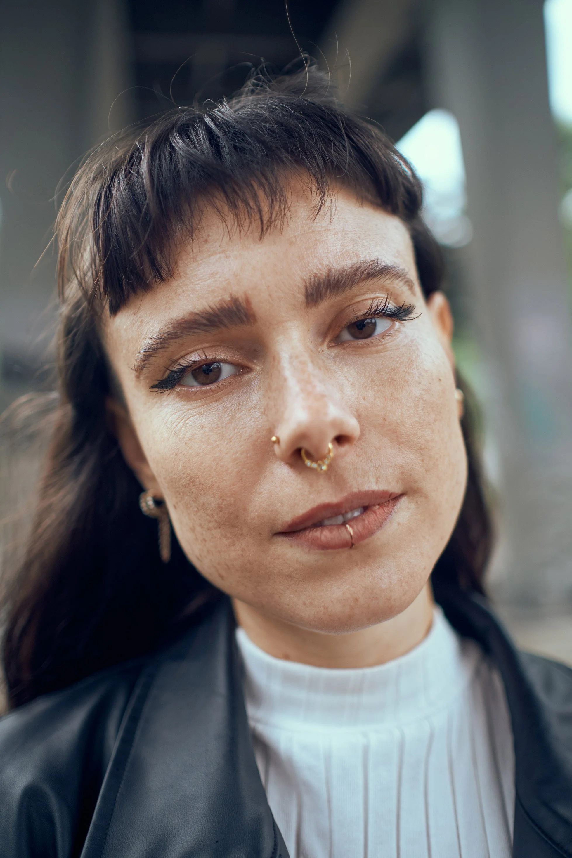 Nahaufnahme eines jungen Frau mit dunklen Haaren, Piercings und natürlichem Make-up, trägt eine schwarze Jacke und einen weißen hochgekrempelten Hemd, im Hintergrund verwaschenes urbane Umgebung.