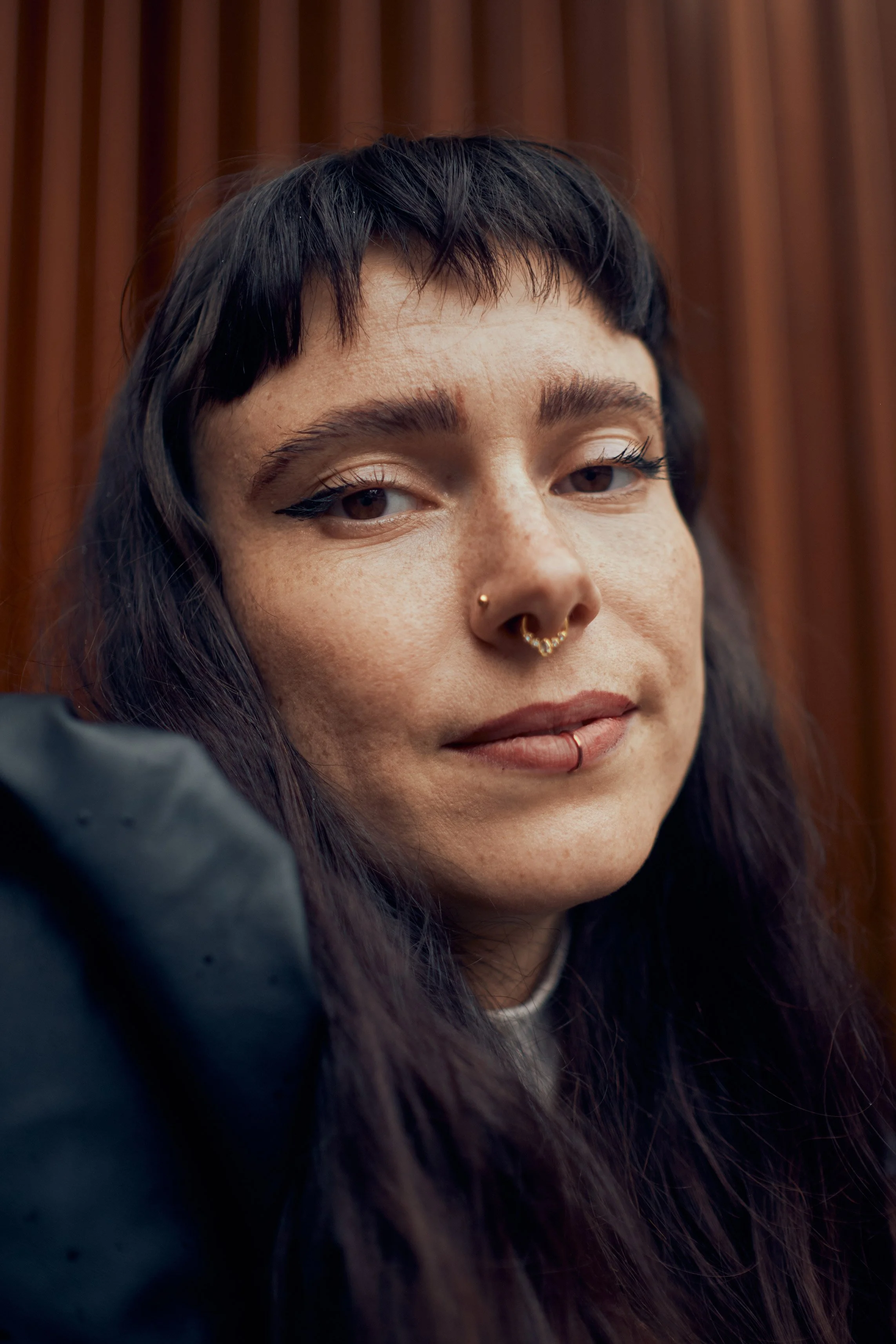 Nahaufnahme eines jungen Mädchens mit schwarzen, kurzen, gestuften Haaren, mehreren Piercings im Gesicht, dunkler Kleidung und einem selbstsicheren Ausdruck, vor einem Holzhintergrund.