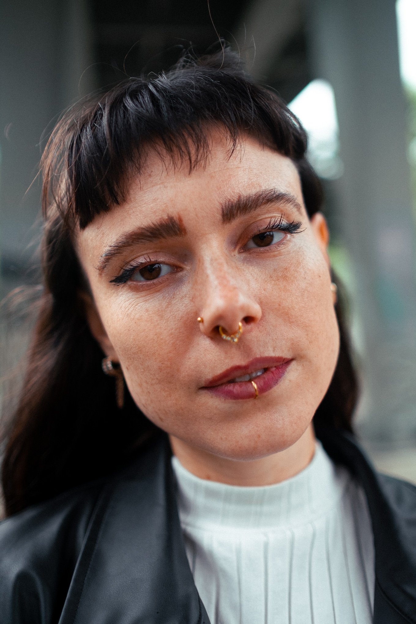 Nahaufnahme einer Frau mit dunklem kurzem Haar, Piercings im Gesicht, dunkler Kleidung und natürlichem Make-up.