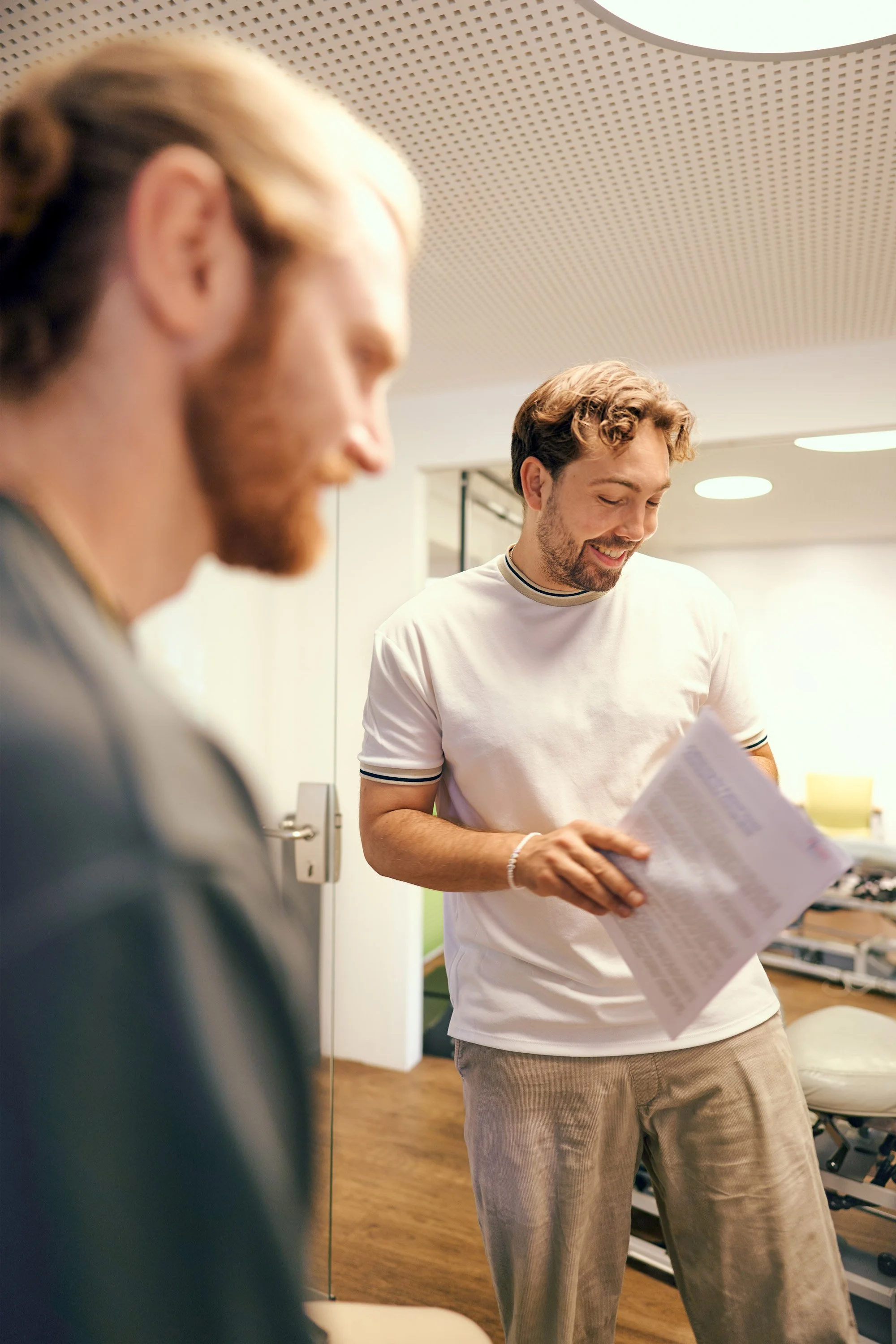 Zwei Männer lachen und schauen auf ein Dokument in einem hell erleuchteten Raum, vermutlich im Büro oder in einer Praxis, mit Holzfußboden und moderner Decke.