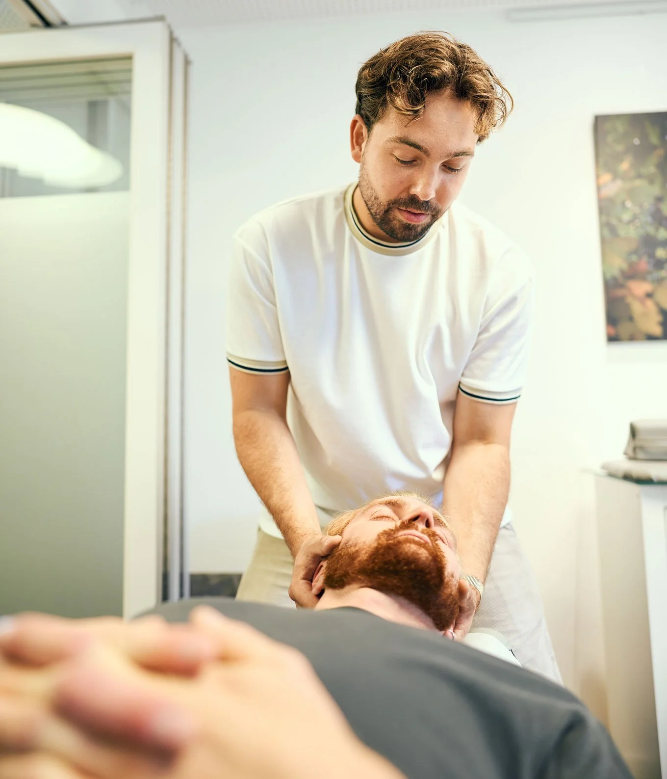 Ein Mann im weißen T-Shirt gibt eine medizinische Behandlung an einem anderen Mann mit Bart, der auf einer Behandlungsliege liegt.