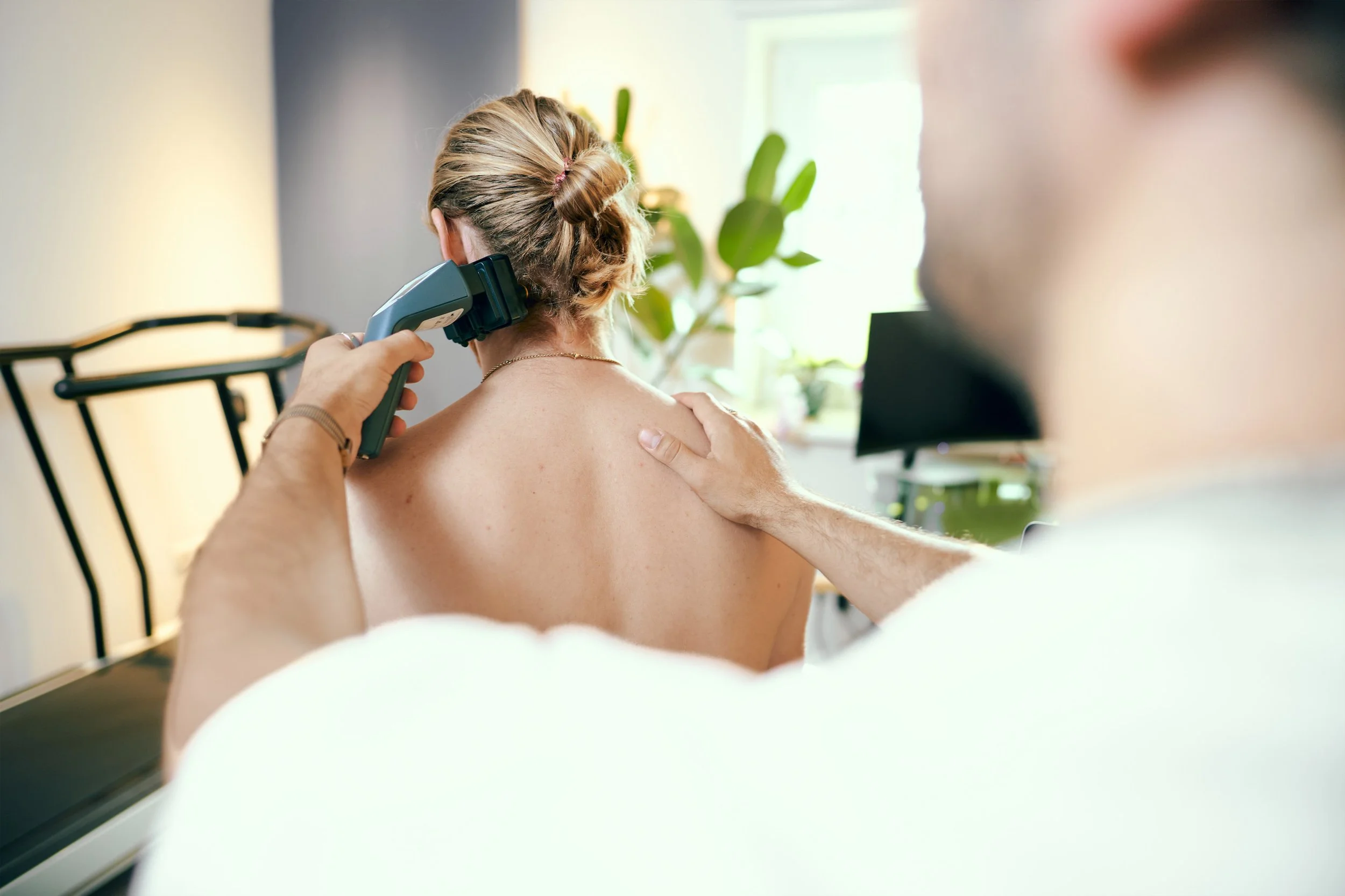 Ärztin führt eine medizinische Untersuchung am Rücken einer Patientin durch, während ein Mann die Patientin berührt. Natürliche Beleuchtung durch Fenster, Pflanzen im Hintergrund.