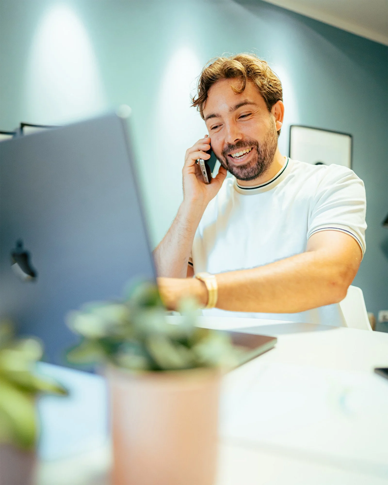 Lächelnder Mann spricht am Telefon während er an einem Laptop arbeitet in einem modernen Raum.