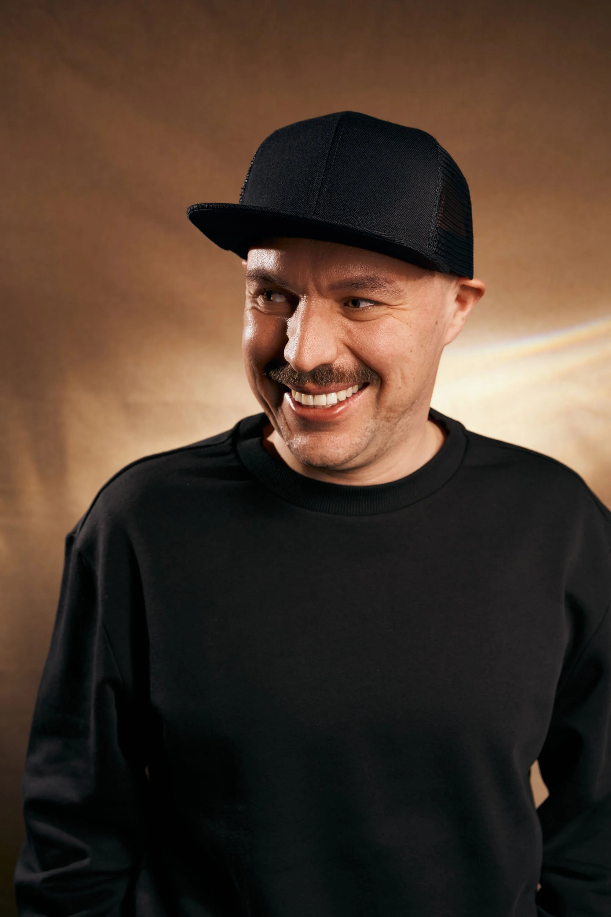 Ein lachender Mann trägt eine schwarze Mütze und ein schwarzes Sweatshirt vor einem braunen Hintergrund