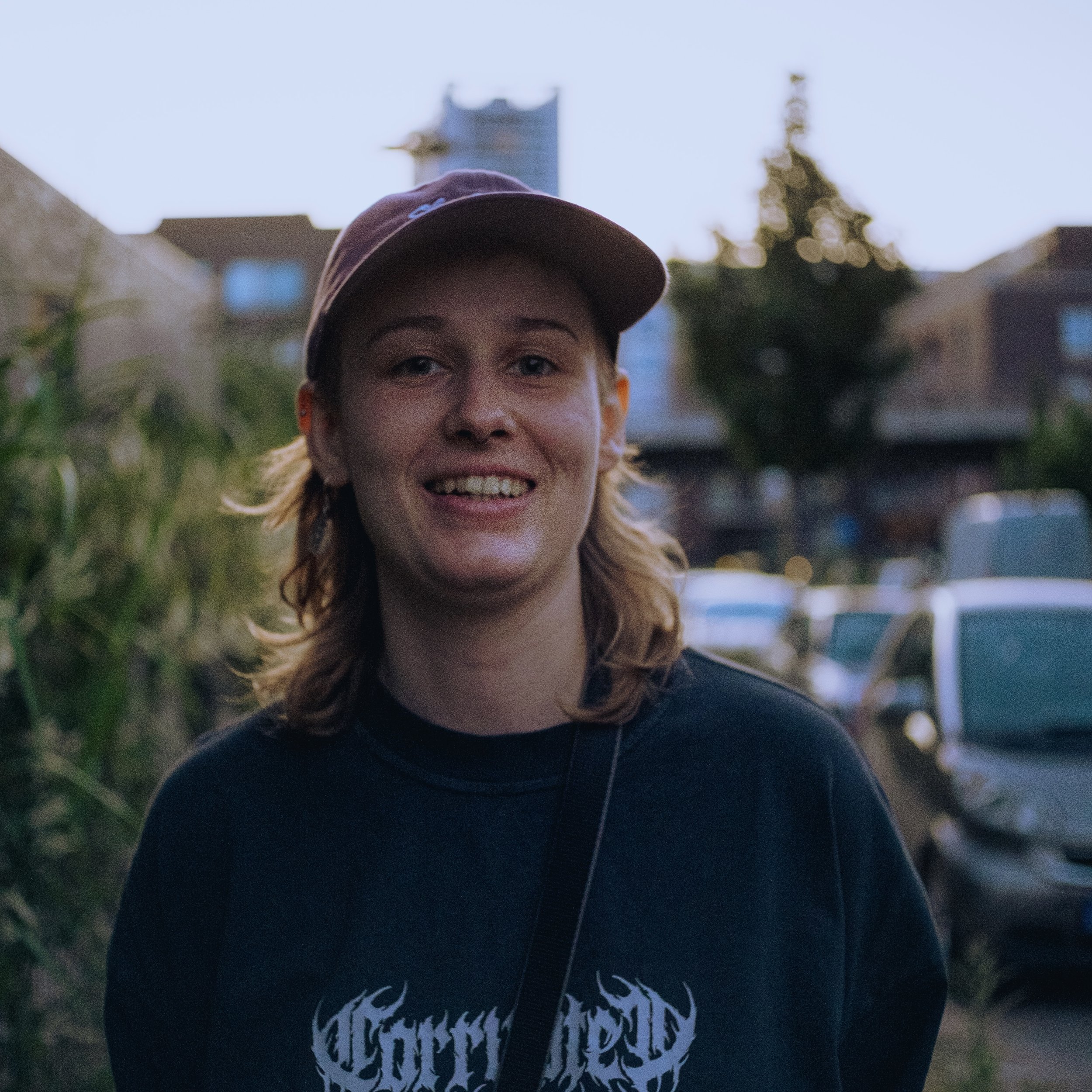 Junge Frau mit braunem Baseballkappe und schwarzem T-Shirt, lächelnd im Freien bei Sonnenuntergang, Hintergründe mit Häusern und Autos