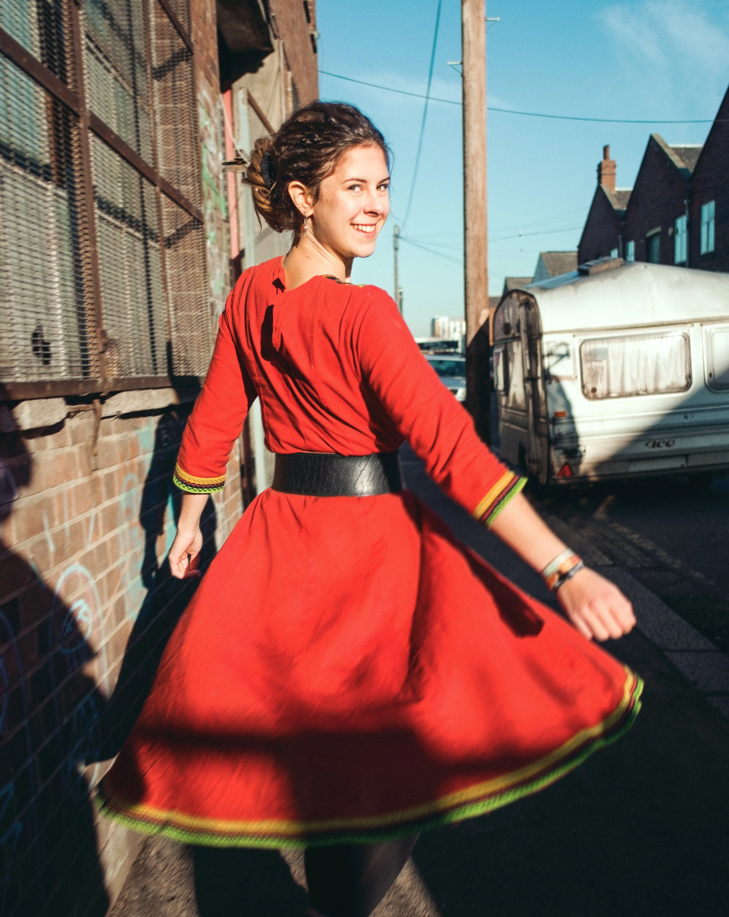 Eine junge Frau in rotblauem Kleid, die auf einer Straße in der Sonne tanzt und lächelt.