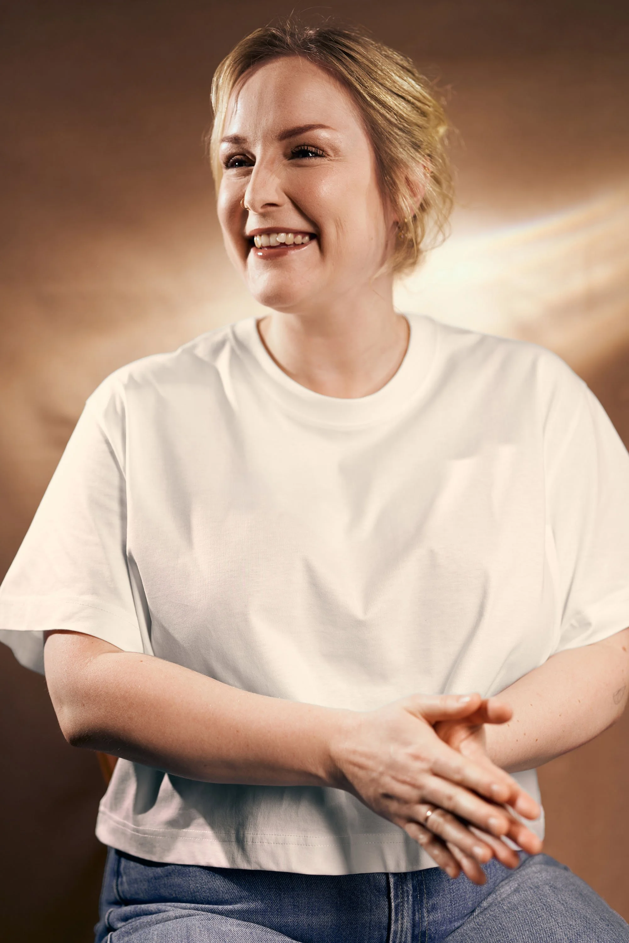 Lächelnde junge Frau mit kurzer brauner Frisur und weißen T-Shirt vor braunem Hintergrund.