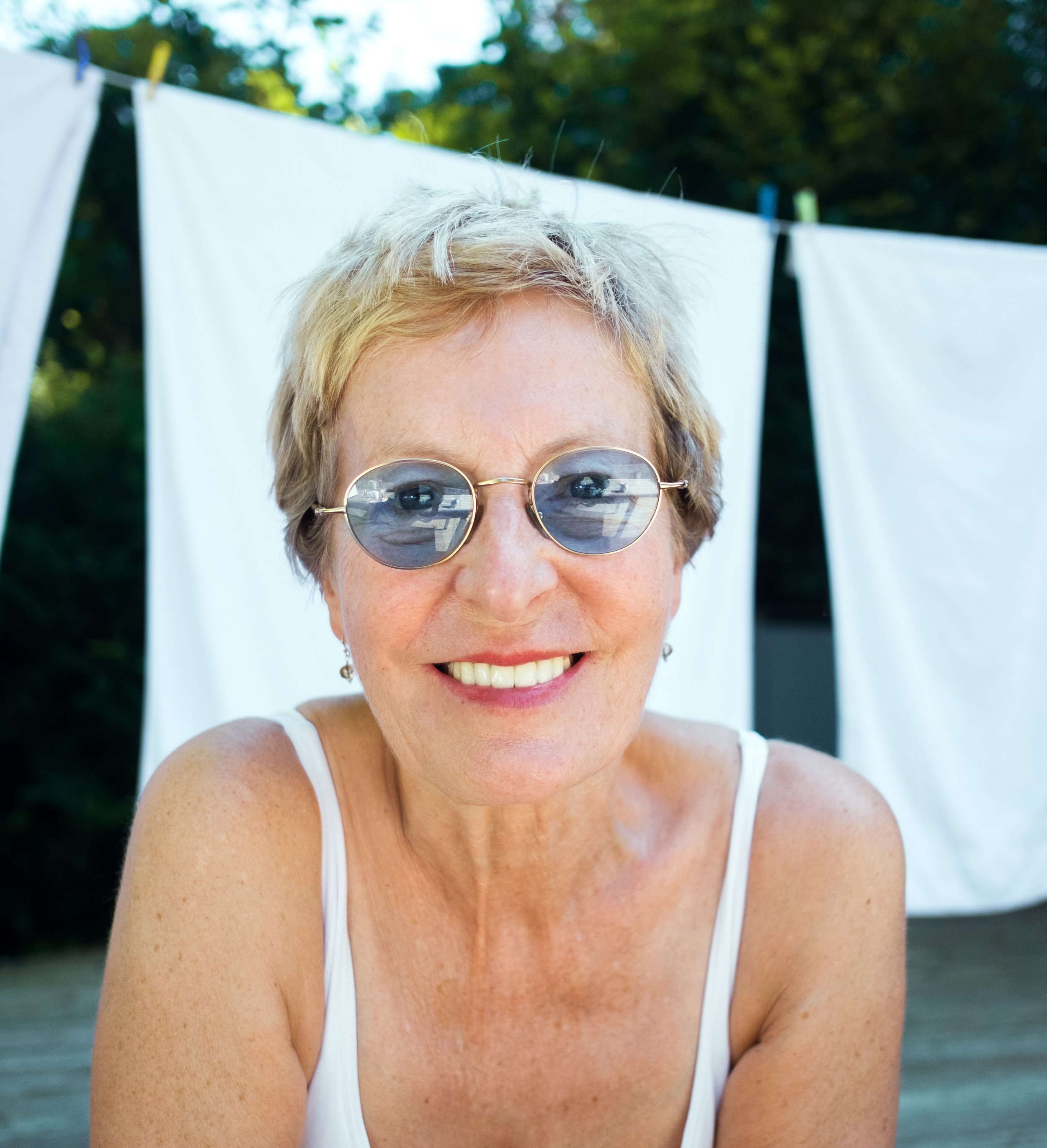 Eine lachende ältere Frau mit kurz blondem Haar, Sonnenbrille und weißem Tank-Top im Freien vor einer Wäscheleine mit weißen Kleidern oder Tüchern.