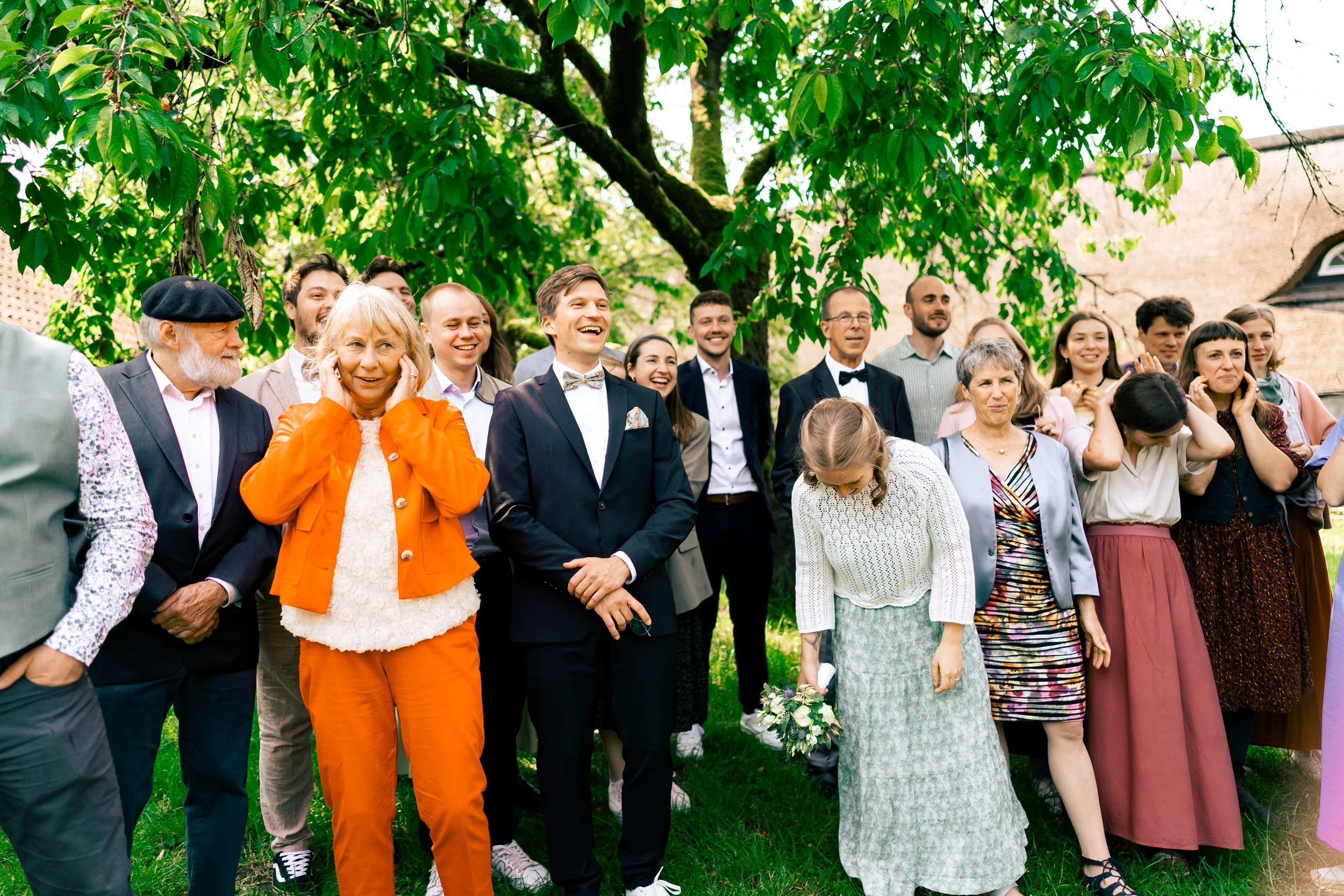 Gruppe von Menschen im Garten unter einem großen Baum bei einer Feier, die meisten lachen oder sind fröhlich, einige Frauen und Männer tragen elegante Kleidung.