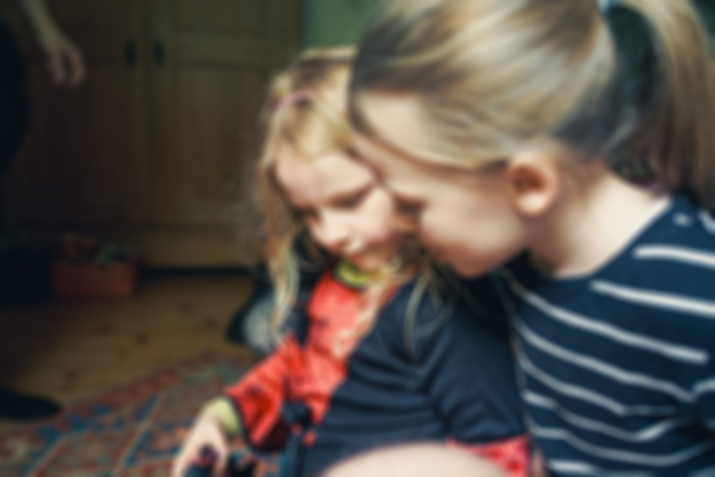 Zwei Kinder sitzen zusammen auf einem Teppich im Wohnzimmer und schauen auf etwas in der Nähe, beide lachen und wirken glücklich.