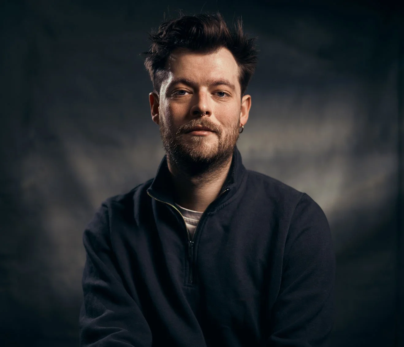 Porträt eines Mannes mit dunklem Haar, Bart und Ohrring, der eine schwarze Jacke trägt, vor dunklem Hintergrund.
