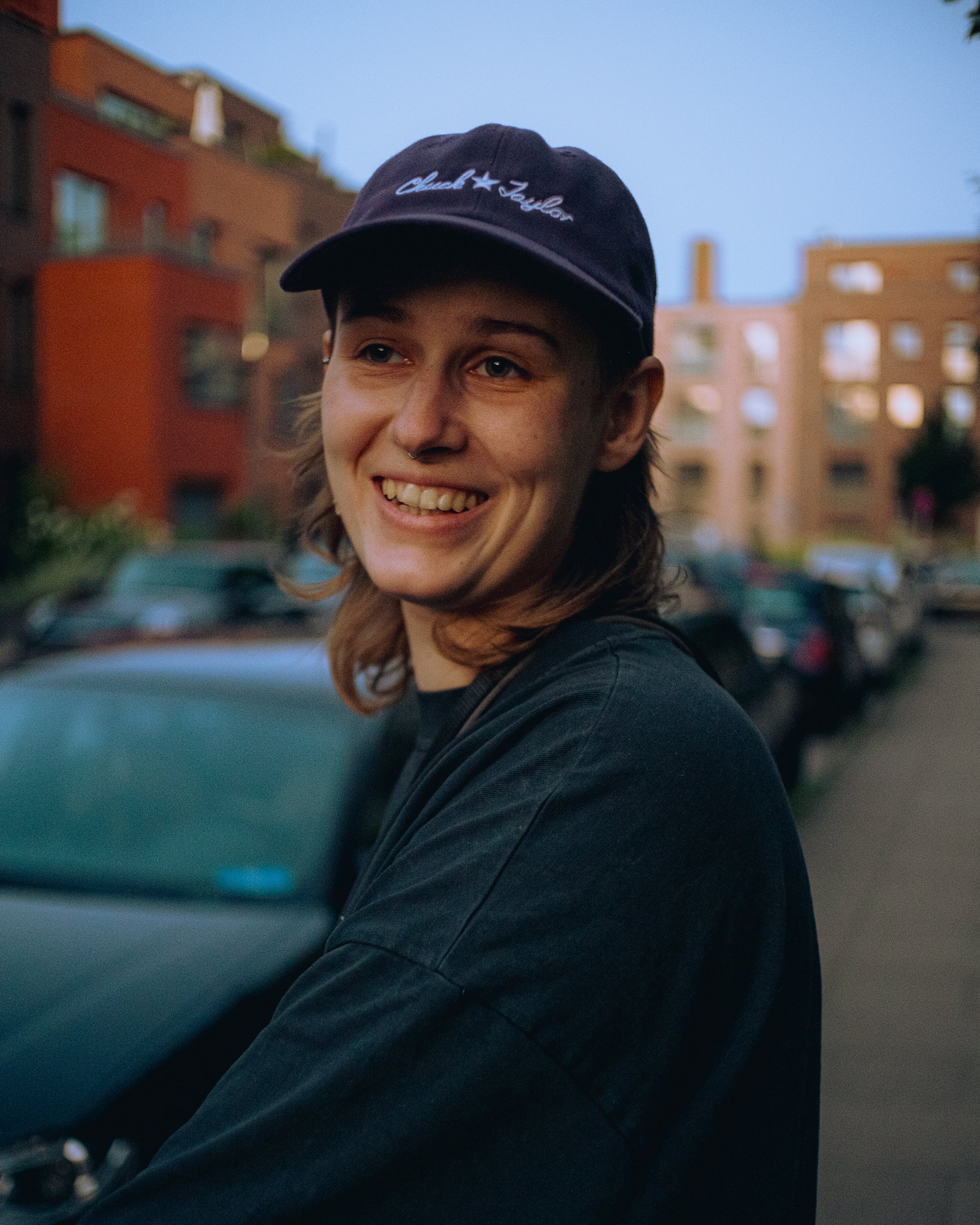 Junge Frau mit braunen Haaren, schwarzem T-Shirt und schwarzer Kappe, die auf einer Stadtstraße im Abendlicht lächelnd in die Kamera schaut.