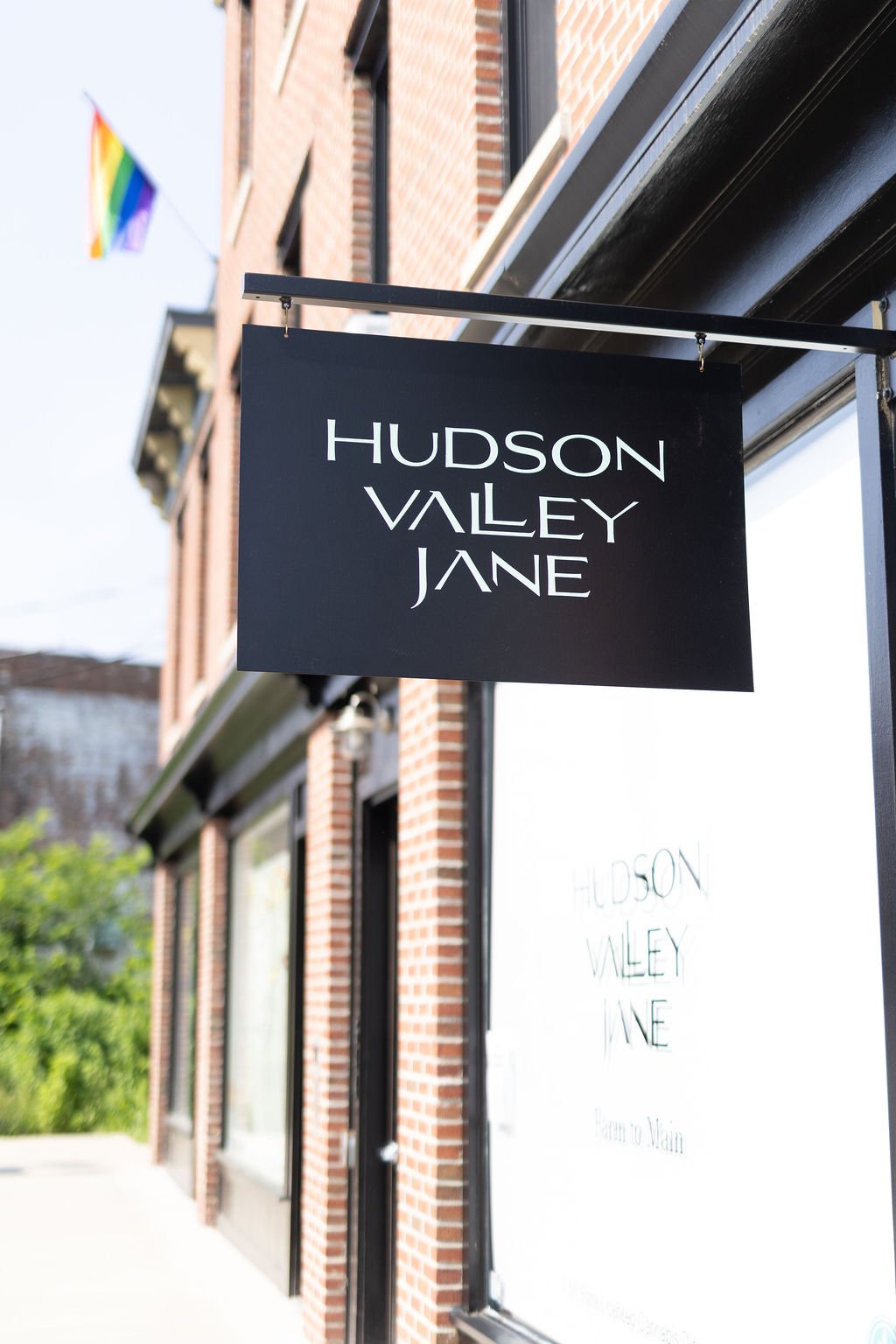 A black sign hanging outside a brick building reads "Hudson Valley Jane" in white letters, with a rainbow flag visible in the background.