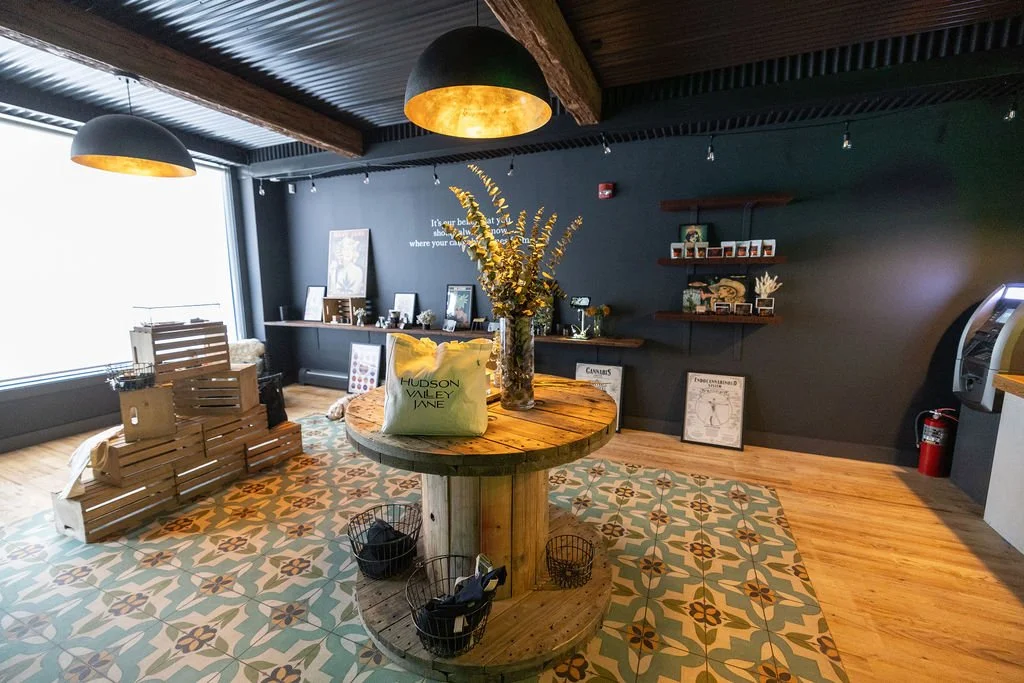 Interior of a cozy shop or cafe with a wooden round table in the center holding a vase with tall yellow flowers and a tote bag. The floor features colorful patterned tiles, and there are wooden crates to the left, framed pictures and decorations on a counter in the background, and dark walls with floating shelves displaying small items. Two black pendant lights hang from the ceiling.