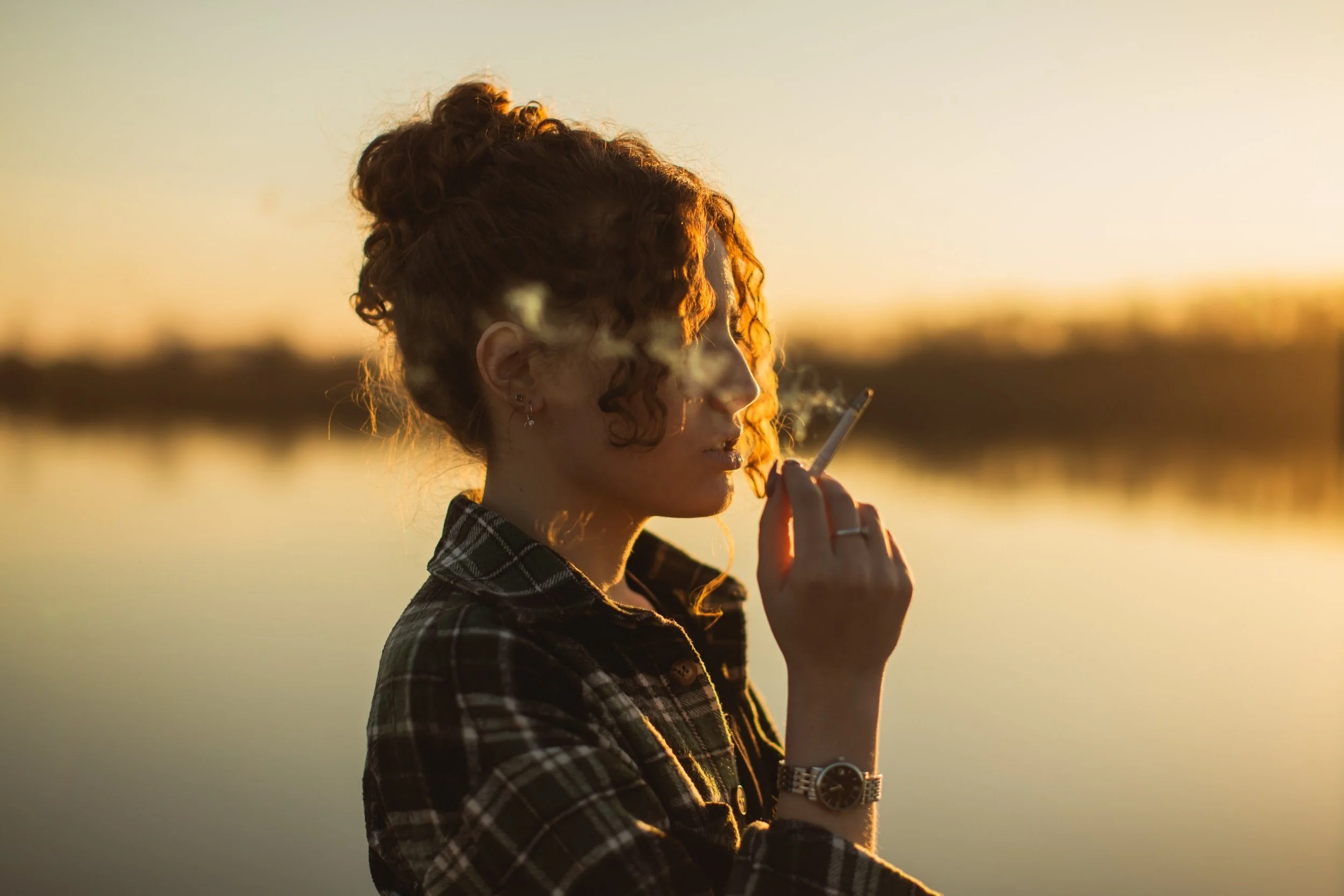 A woman with curly hair wearing a plaid shirt and watch, smoking a cigarette at sunset by a body of water.